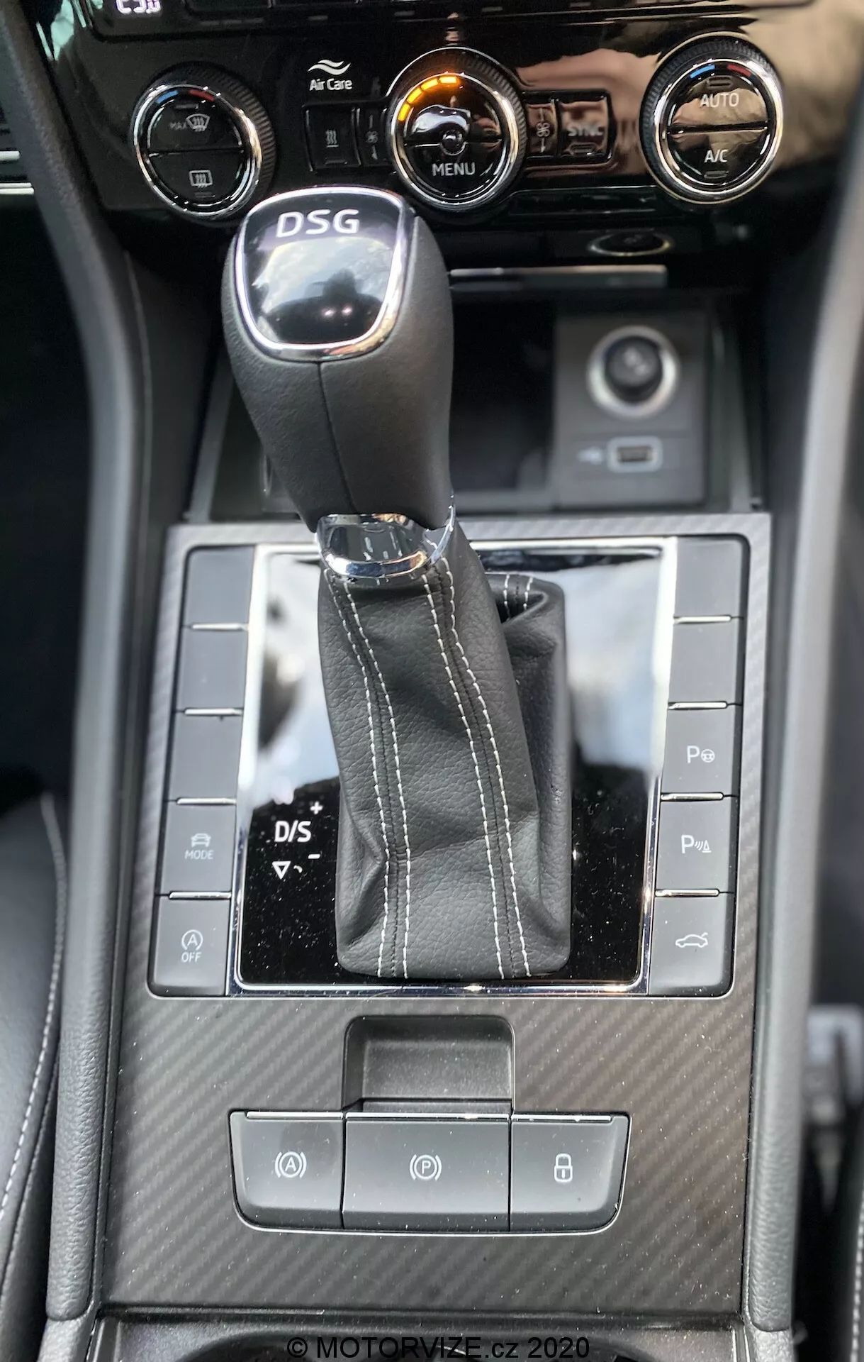Vista interna della console centrale della Skoda Superb 2019 da un angolo discendente, evidenziando la leva del cambio DSG, pulsanti di controllo simmetrici, dettagli testurizzati simili alla fibra di carbonio e comandi del clima con touchscreen centrale che mostra informazioni sul veicolo.