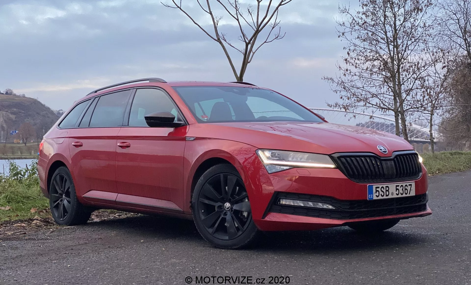 Immagine di una Škoda Superb station wagon rossa con vista anteriore tre quarti, che mostra il design distintivo della griglia con il logo Škoda, fari a LED, cofano scolpito, luci fendinebbia, cerchi in lega neri a più razze, finestrini oscurati e specchietti laterali con indicatori di direzione integrati, posizionati in un ambiente rurale tranquillo con fogliame e acqua sullo sfondo.