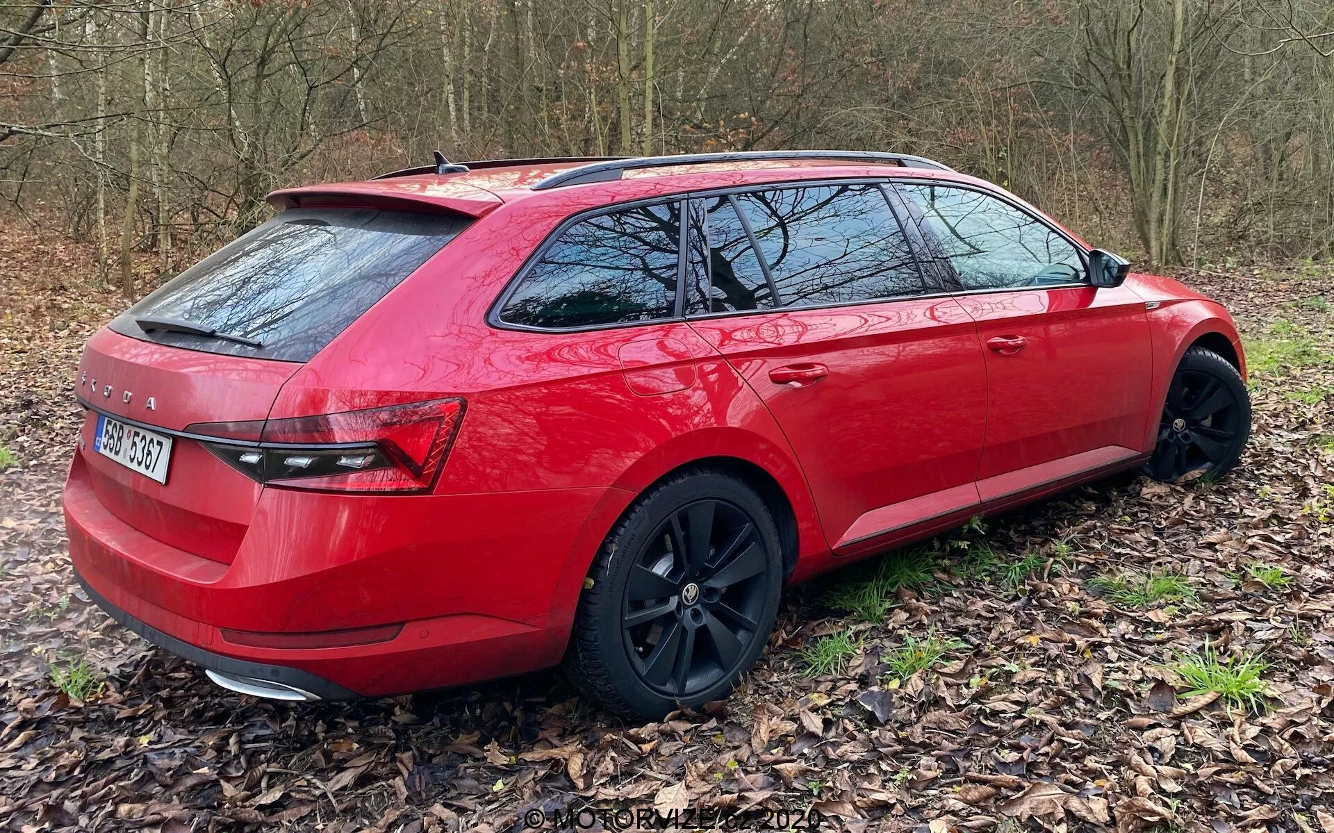 Una station wagon rossa vista da una prospettiva posteriore di tre quarti, posizionata fuori strada su un letto di foglie. Si cattura il lato destro del veicolo, dal paraurti posteriore alle porte anteriori, mostrando il suo design moderno e premium. I cerchi in lega neri sono leggermente girati a destra, aggiungendo al suo aspetto sportivo e di alta gamma. Le caratteristiche includono gruppi ottici posteriori eleganti, pneumatici a basso profilo e un sistema di barre sul tetto. I finestrini posteriori oscurati suggeriscono maggiore privacy e protezione solare.