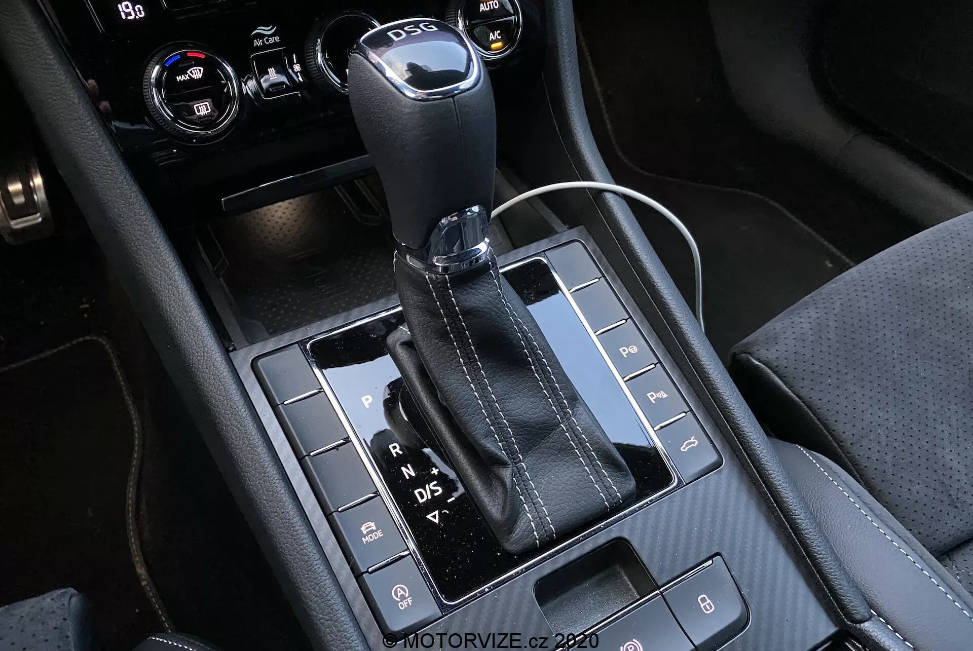 Vista ravvicinata della leva del cambio automatico e dell'area della console centrale della Skoda Superb 2019, con una leva del cambio rivestita in pelle con parte superiore in metallo e lettere visibili 'P, R, N, D/S' che indicano le diverse marce. Il pannello nero lucido ospita vari pulsanti per funzionalità come sensori di parcheggio, disattivazione del sistema Auto Start/Stop e controllo della trazione. La console mostra una combinazione di materiali con cuciture dettagliate, inclusa una presa di corrente da 12 volt e probabilmente comandi per il clima o l'infotainment nell'angolo in alto a destra ritagliato.