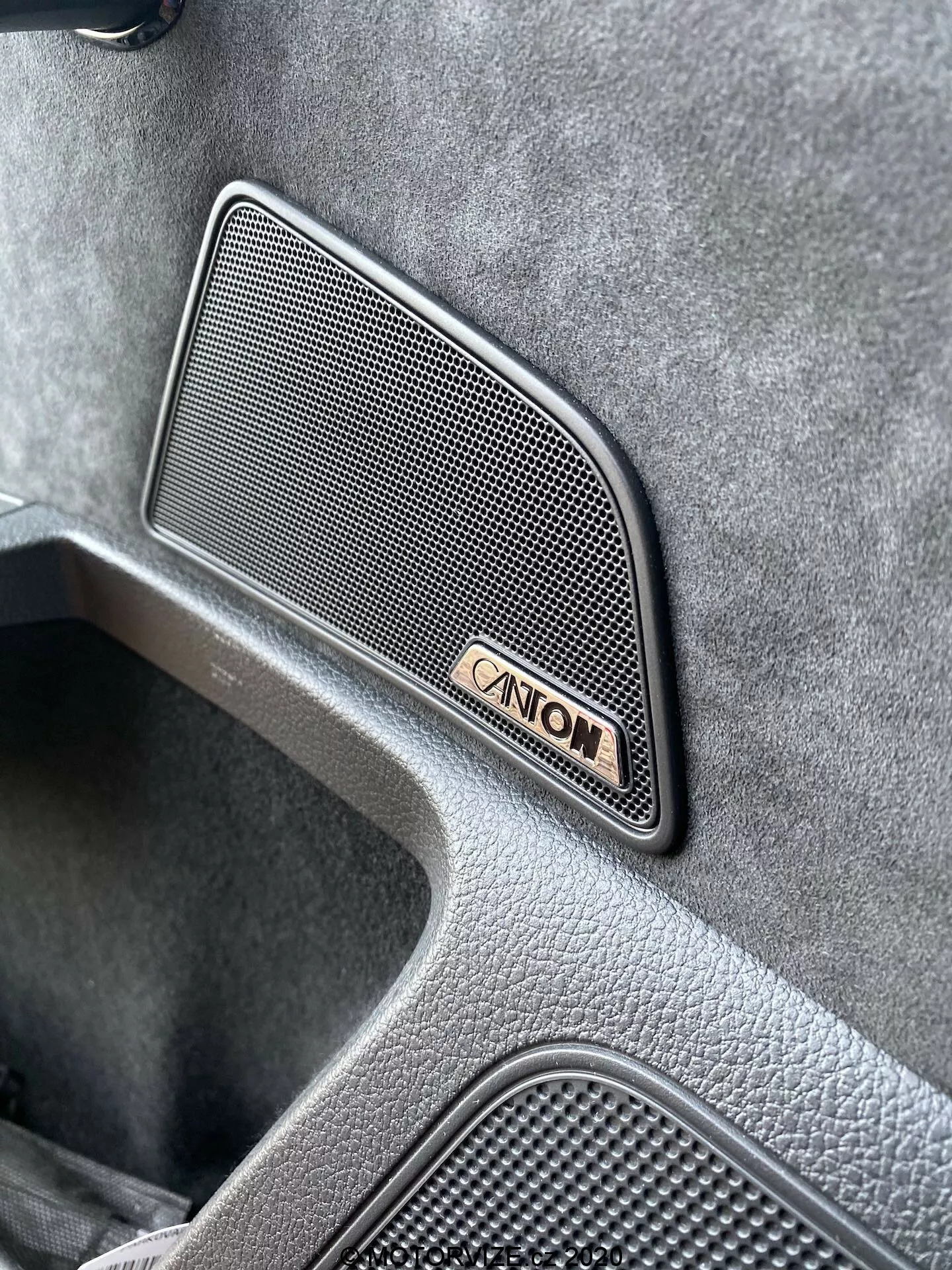 Vista angolata ravvicinata degli interni della Skoda Superb Limousine 2019, evidenziando una griglia altoparlante audio Canton premium con forma trapezoidale a motivo a rete, situata sul pannello della porta, accompagnata dall'emblema Canton, mostrando una finitura testurizzata di alta qualità e un'area incassata indicativa di un design veicolare di lusso.