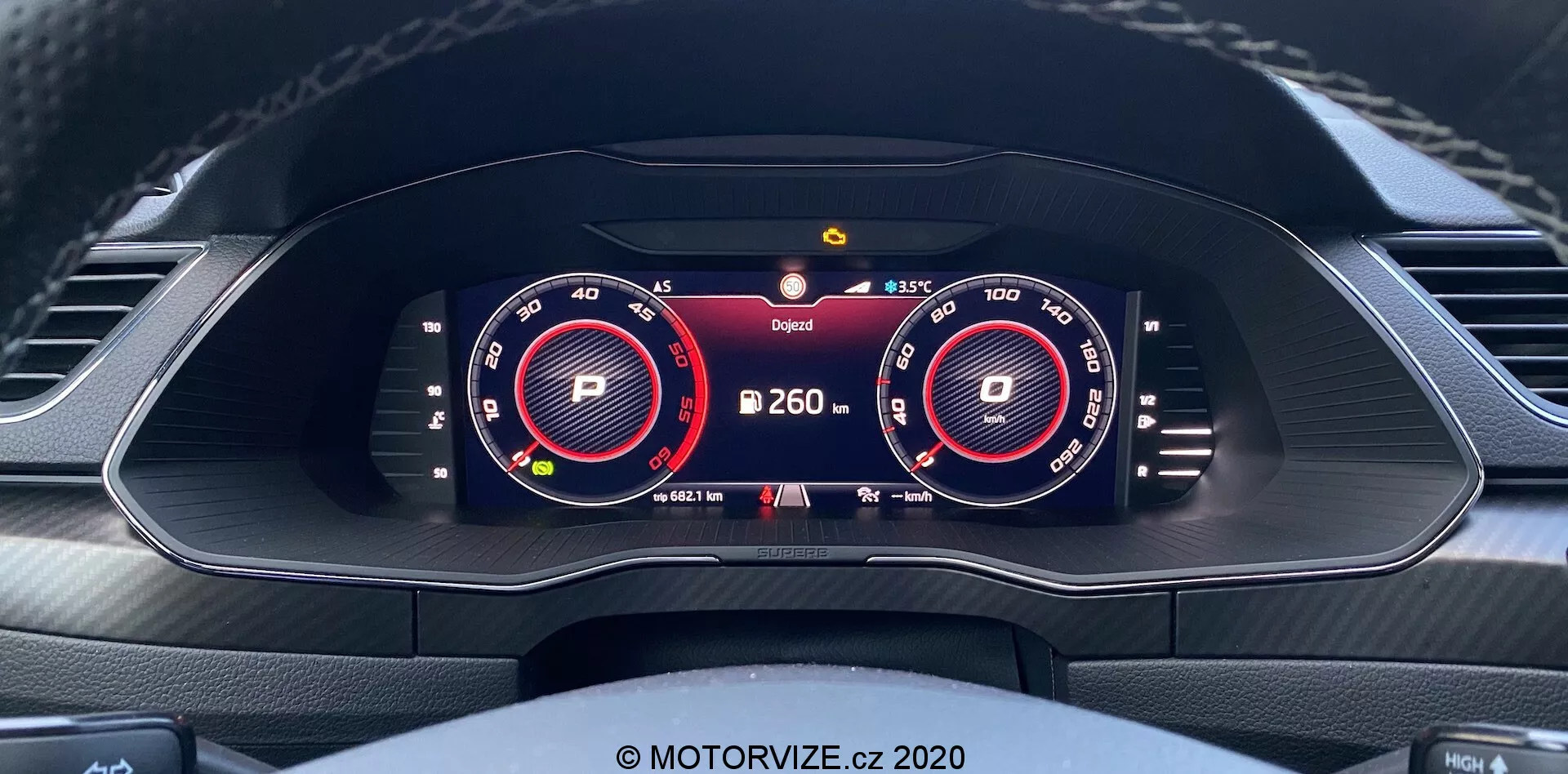 Vista interna della Škoda Superb Limousine 2019, focalizzata su un quadro strumenti completamente digitale e ad alta risoluzione dalla prospettiva del conducente. Il display include due grandi quadranti virtuali per il tachimetro e il tachimetro, illuminati in rosso e bianco. Tra i quadranti vengono visualizzate informazioni aggiuntive, come l'indicatore del cambio 'P', la lettura della temperatura e un simbolo di avvertimento. Un indicatore della batteria suggerisce capacità elettriche o ibride. Il design del cruscotto include prese d'aria e la parte superiore di un volante sagomato con dettagli di cucitura.