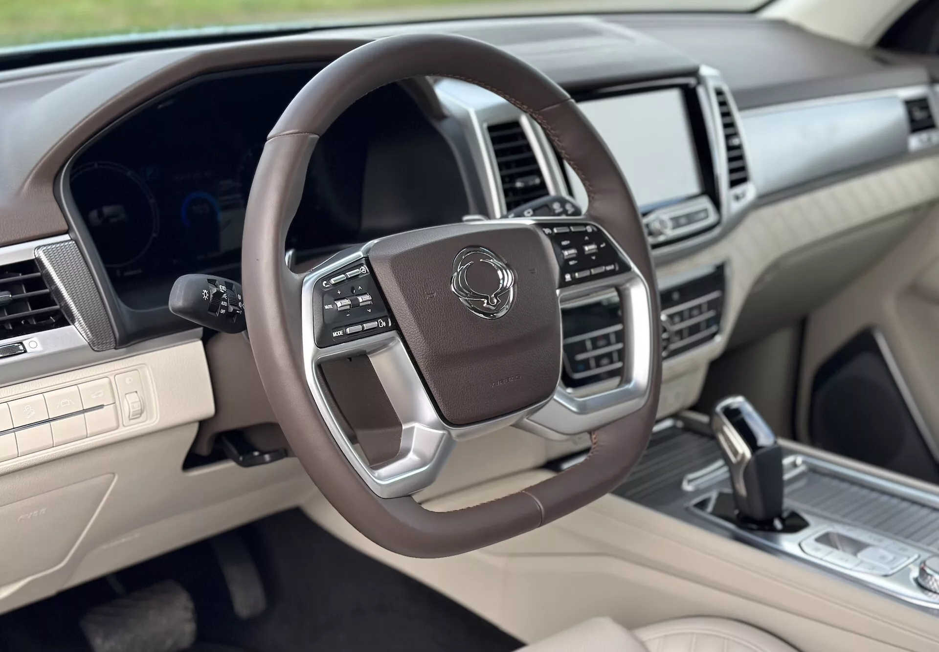 Vista dell'interno lussuoso della SsangYong Rexton 2021 dal lato del passeggero anteriore, con volante multifunzione rivestito in pelle con accenti metallici, cluster strumenti digitale, sistema infotainment avanzato, e schema bicolore beige e scuro.