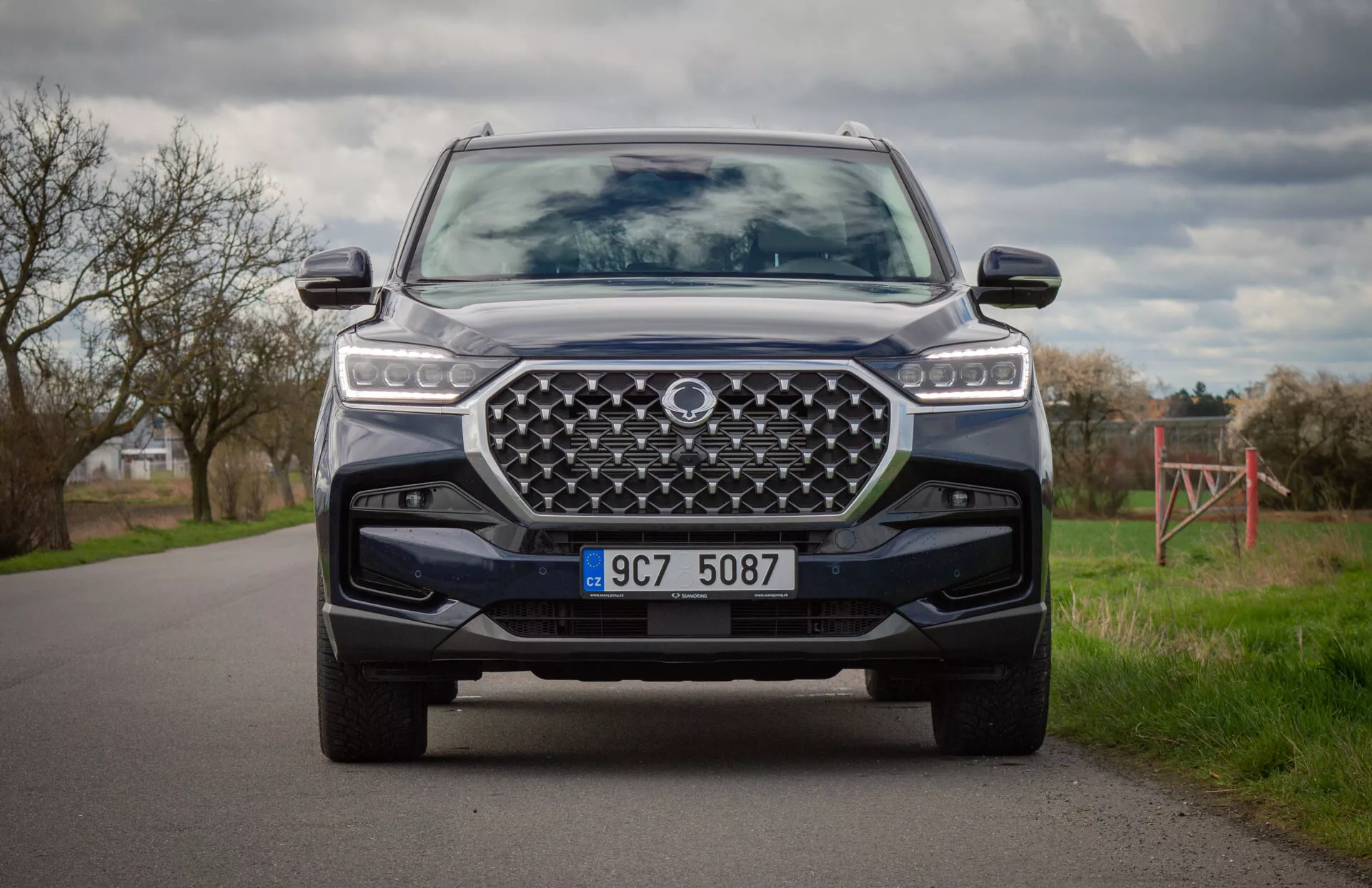SUV moderno SsangYong Rexton 2021 a colori, ripreso da una posizione inginocchiata che mostra il frontale del veicolo, fari LED, grande griglia frontale disegnata, paraurti anteriore con piastra di protezione e sistemi avanzati di assistenza al guidatore
