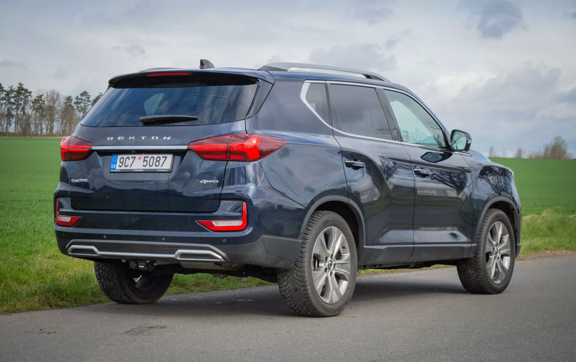 SUV SsangYong Rexton di colore scuro parcheggiato su una superficie asfaltata con fanali posteriori LED, riflettori posteriori di sicurezza con sensori di parcheggio, ruote in lega, vetri con privacy tinteggiati, barre sul tetto, antenna a pinna di squalo, tubo di scarico visibile, altezza da terra elevata e segnali di svolta integrati negli specchietti laterali su sfondo di campo erboso e cielo coperto.