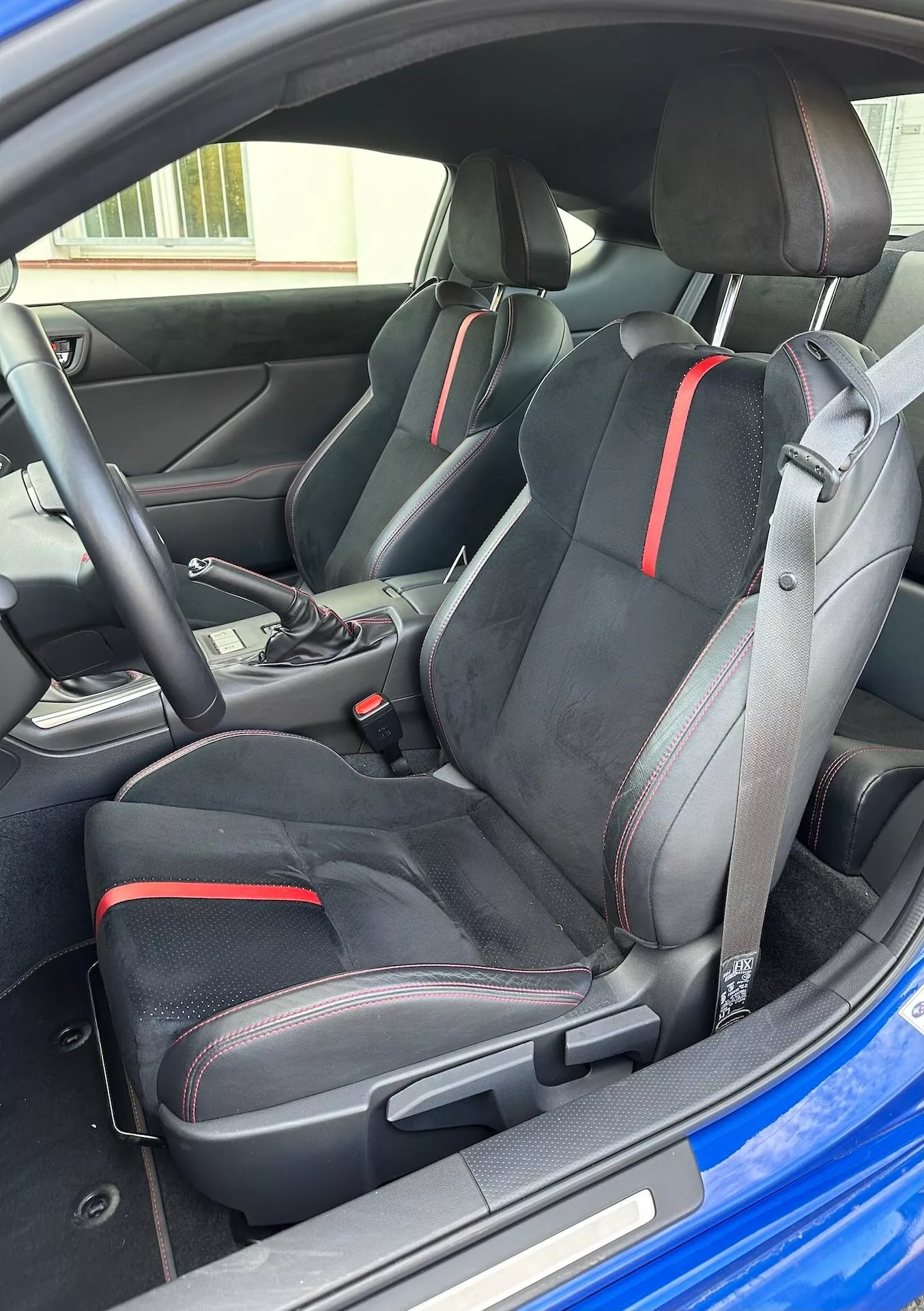 Vista interna della Subaru BRZ 2023 che evidenzia i sedili sportivi anteriori con design ergonomico e dettagli rossi, selettore di trasmissione automatica e estetica moderna del cockpit da una prospettiva esterna obliqua