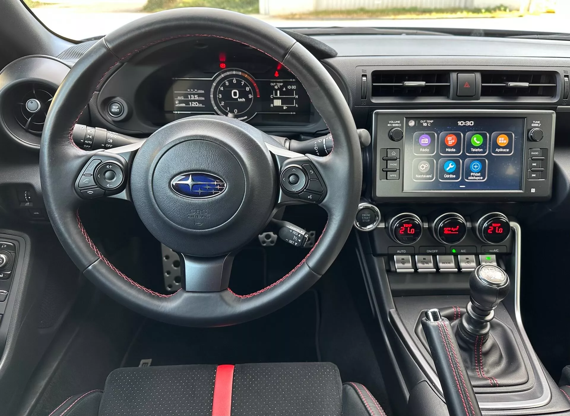 Interno della Subaru BRZ 2023 con prospettiva del guidatore che mostra un volante sportivo in pelle con comandi multifunzione e cuciture rosse, un cluster strumenti con quadranti analogici e display digitale, una consolle centrale con un ampio sistema di infotainment touchscreen e controllo clima bizona, e un cambio manuale con copertura in pelle e accenti rossi, contro un interno nero con accenti rossi sportivi, evidenziando le funzionalità avanzate del veicolo e la tecnologia.