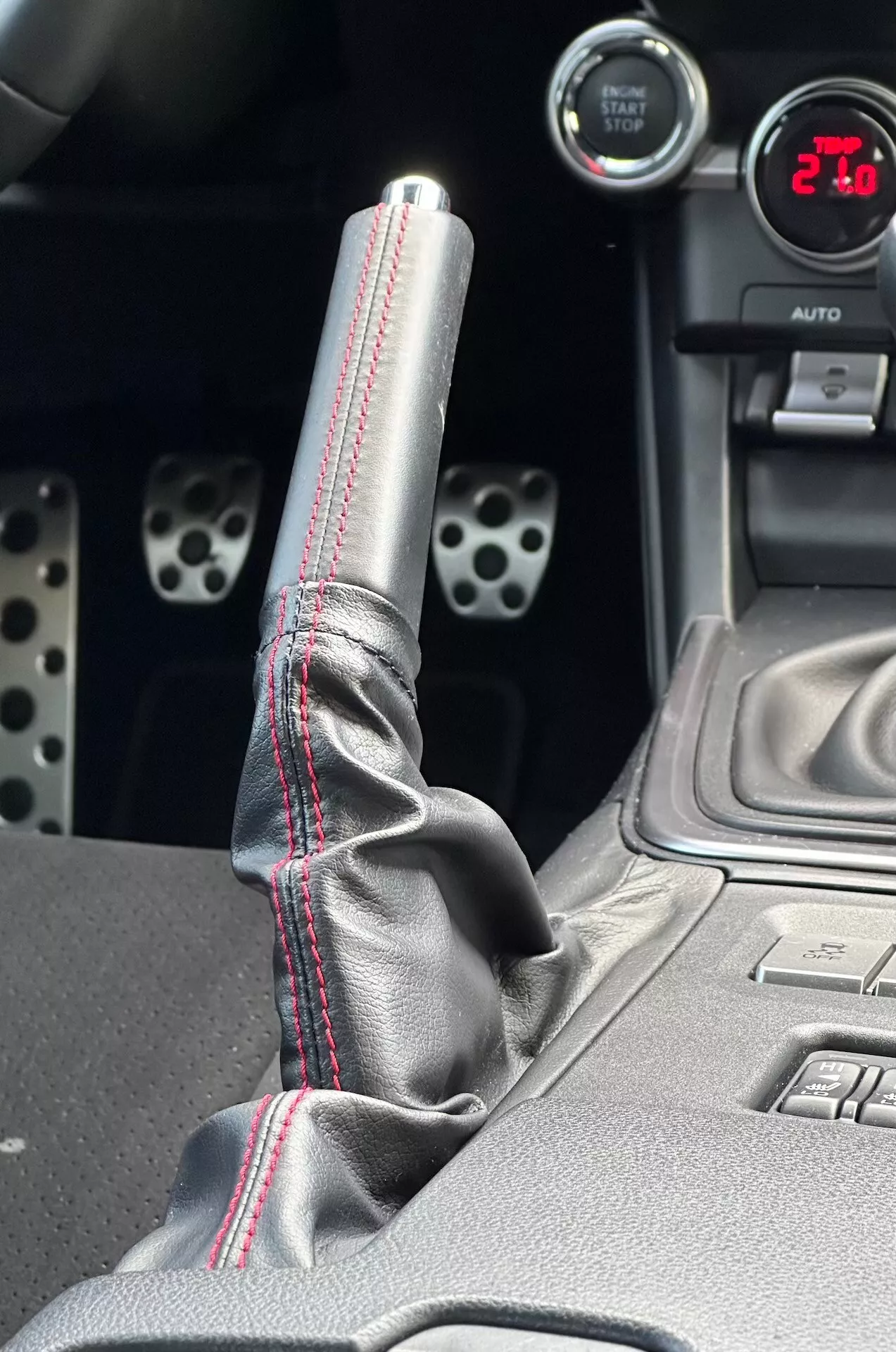 Primo piano della console centrale della Subaru BRZ 2023 mettendo in evidenza la leva del cambio in metallo con cuciture rosse a contrasto sulla copertura in pelle, indicatore digitale che mostra '21', pedali sportivi in alluminio con fori trapanati, e caratteristiche tecnologiche come pulsanti di controllo della stabilità elettronica.