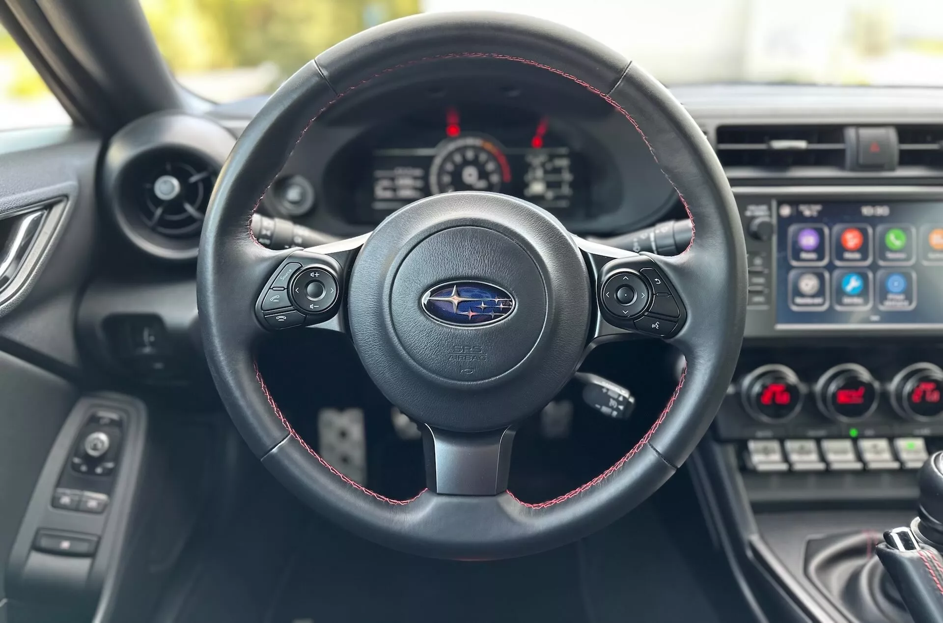 Vista interna del Subaru BRZ 2023 dal sedile del conducente focalizzata sul volante a tre razze con cuciture rosse, logo Subaru e cruscotto con quadro strumenti e console centrale
