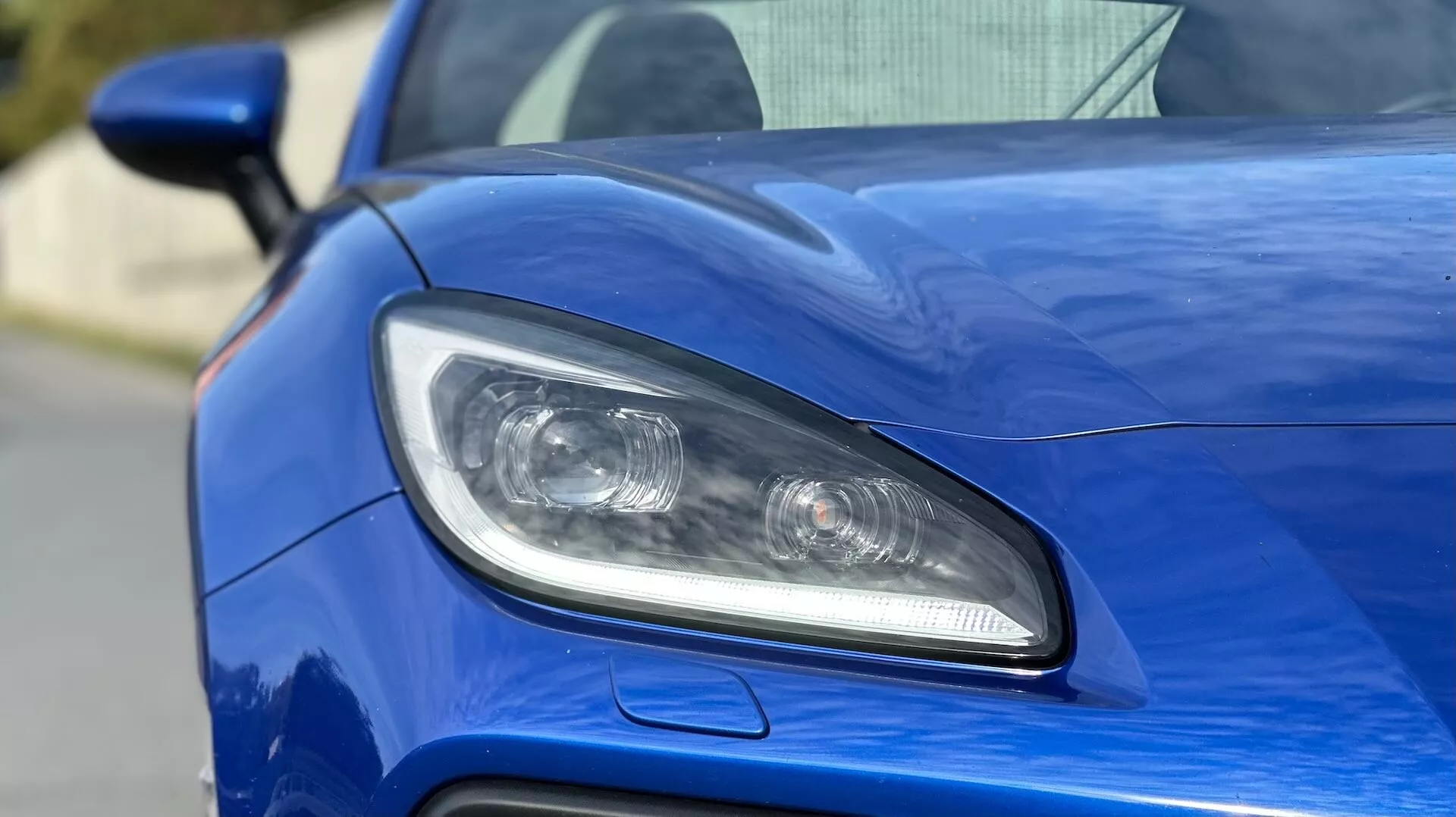 Primo piano del faro lato guidatore della Subaru BRZ 2023, caratterizzato da illuminazione LED o HID, lenti a proiettore, e presa d'aria sportiva su carrozzeria blu metallizzato