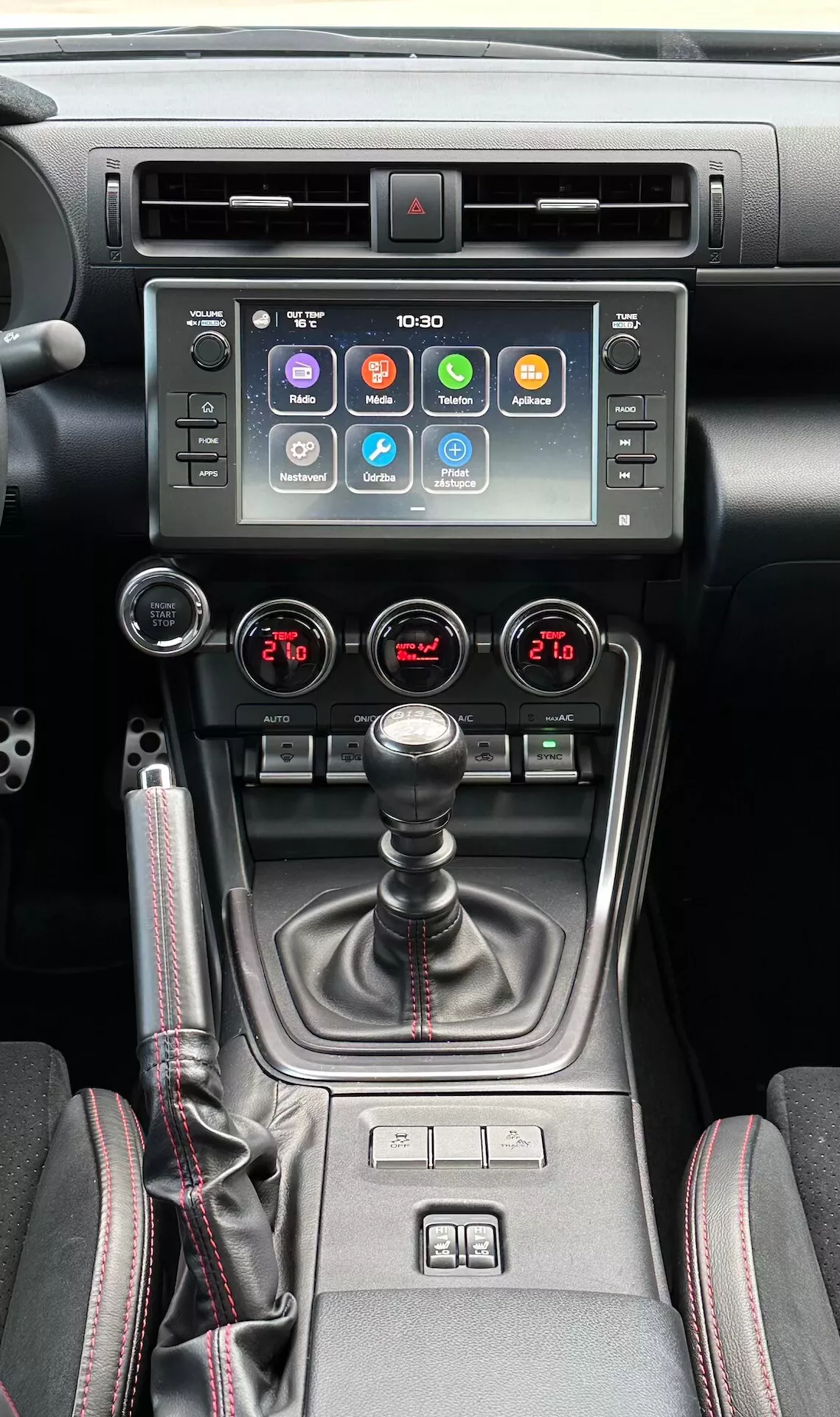 Interno della Subaru BRZ 2023 che mette in mostra la consola centrale, con touchscreen per l'infotainment, indicatore di luce di emergenza, bocchette dell'aria metalliche, controllo del clima a doppia zona, leva del cambio manuale con copertura in pelle, comandi per il riscaldamento dei sedili e un selettore di modalità di guida multipla in un design moderno nero e metallico.