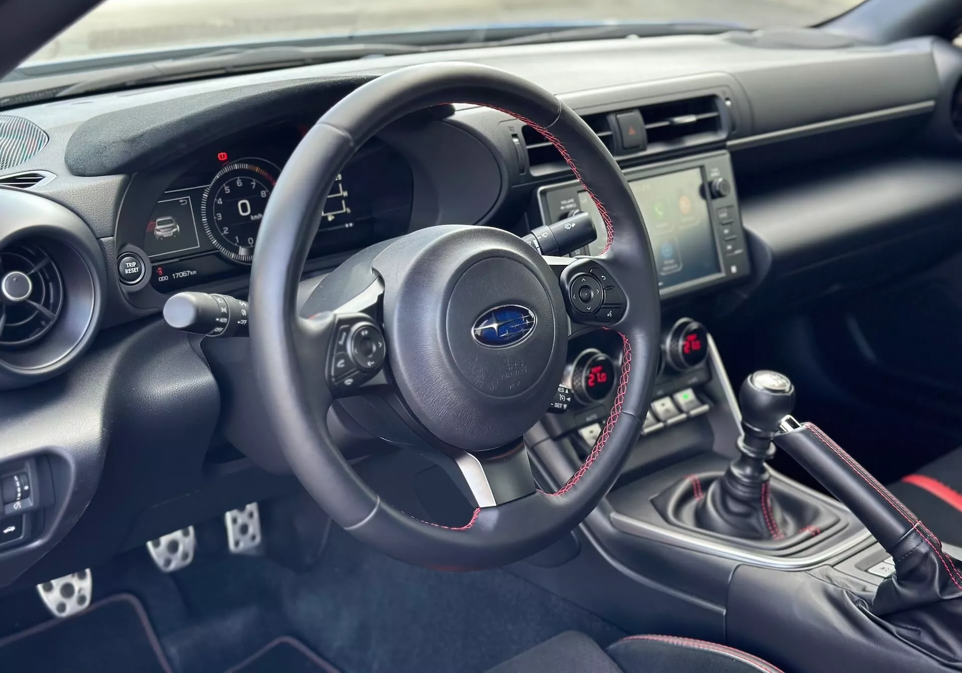 Interno della Subaru BRZ 2023 con focus sul lato guida della plancia e della cabina, vista dettagliata del volante, del quadro strumenti, della consolle centrale con controlli audio e climatizzatore, e leva del cambio manuale con cuciture rosse sportive