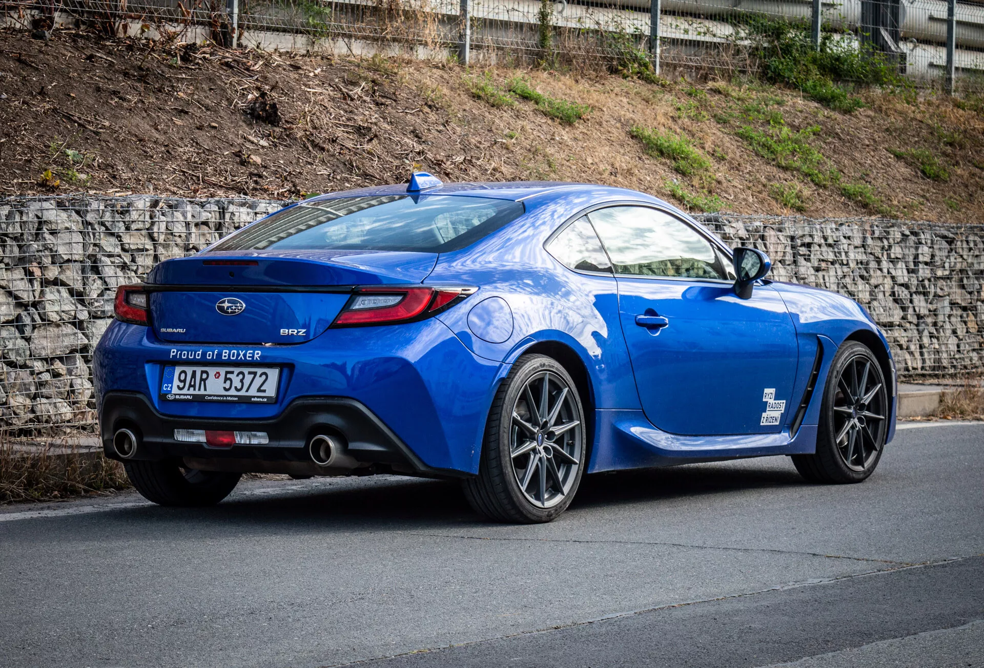 Coupé sportiva Subaru BRZ 2023 in blu metallizzato parcheggiata su una strada in pendenza, mostrando la sua vista posteriore a tre quarti che enfatizza il profilo laterale del veicolo, la curvatura posteriore, le ruote in lega sportive, i terminali di scarico doppi, lo spoiler a labbro e il badge del motore 'Proud of Boxer'.
