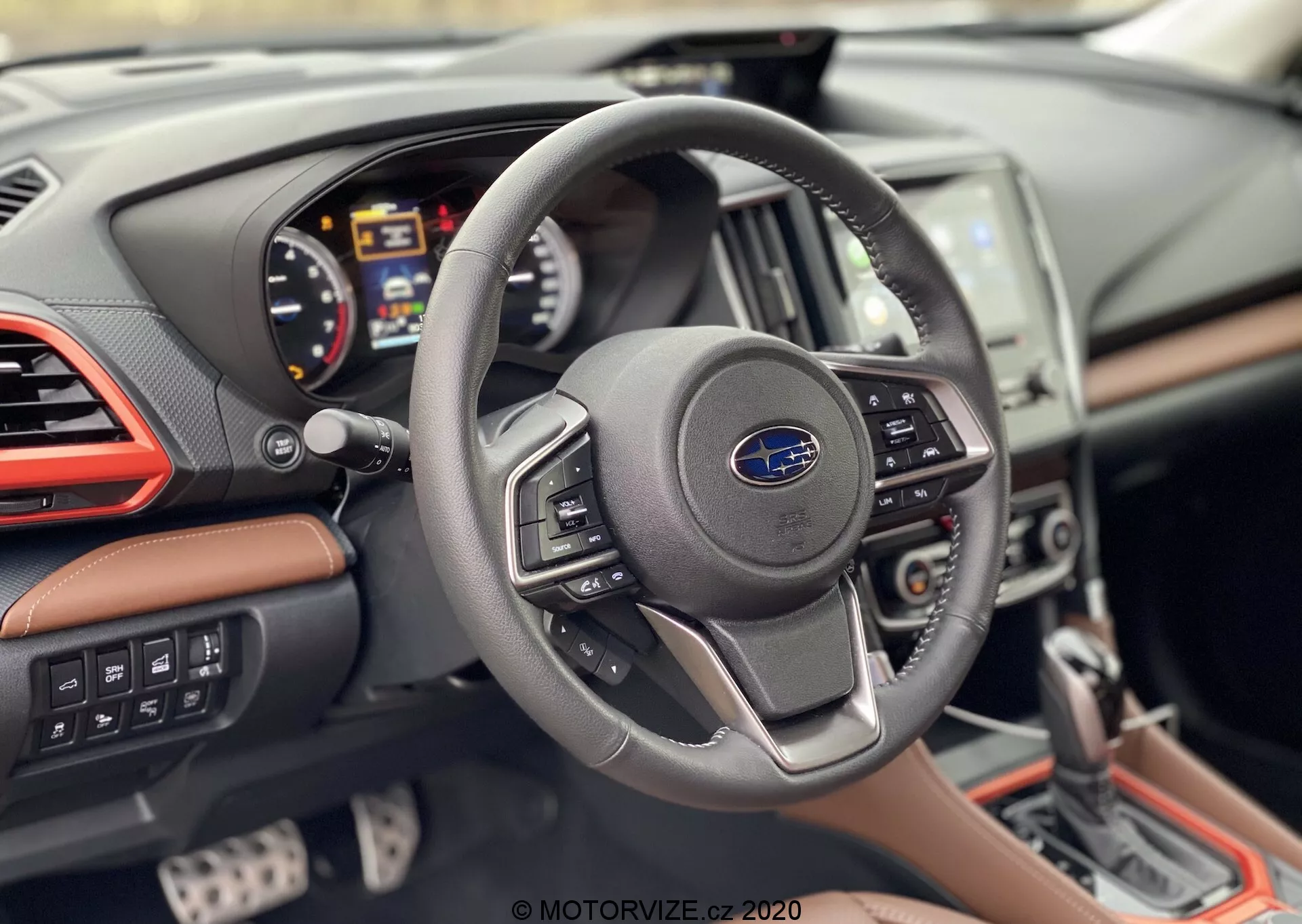 Vista interna della Subaru Forester 2020 focalizzata sulla prospettiva del conducente, mostrando un volante moderno a tre razze con logo Subaru e pulsanti di controllo, quadro strumenti digitale con contagiri, tachimetro e display multi-informazione, console centrale parzialmente visibile con sistema di infotainment touchscreen e cruscotto con rivestimento in pelle, accenti cromati e cuciture a contrasto.