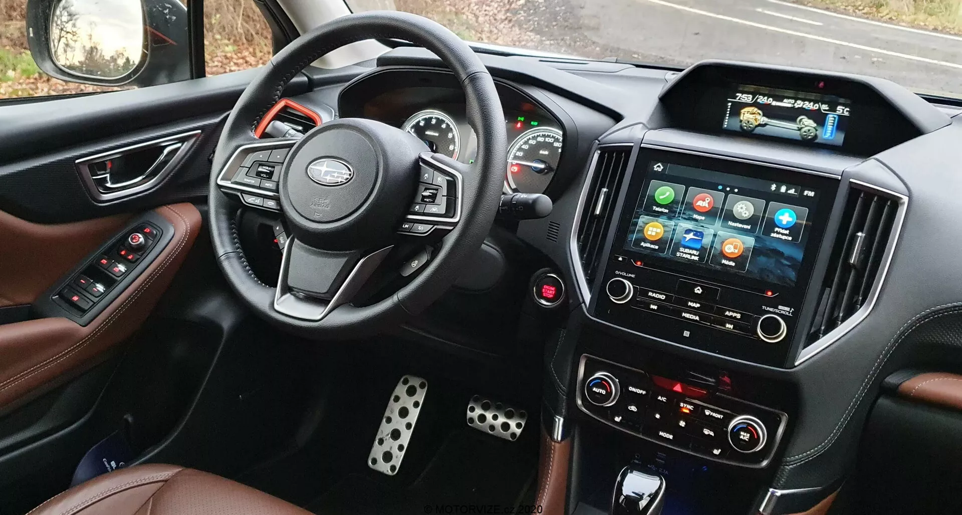 Vista interna del Subaru Forester 2020 dal sedile del conducente, che mostra il volante con controlli montati, il quadro strumenti parzialmente visibile con indicatori analogici e display digitale centrale, il sistema di infotainment touchscreen prominente con icone colorate, pulsanti di controllo del clima manuali, sedili rivestiti in pelle e coperture dei pedali in metallo.