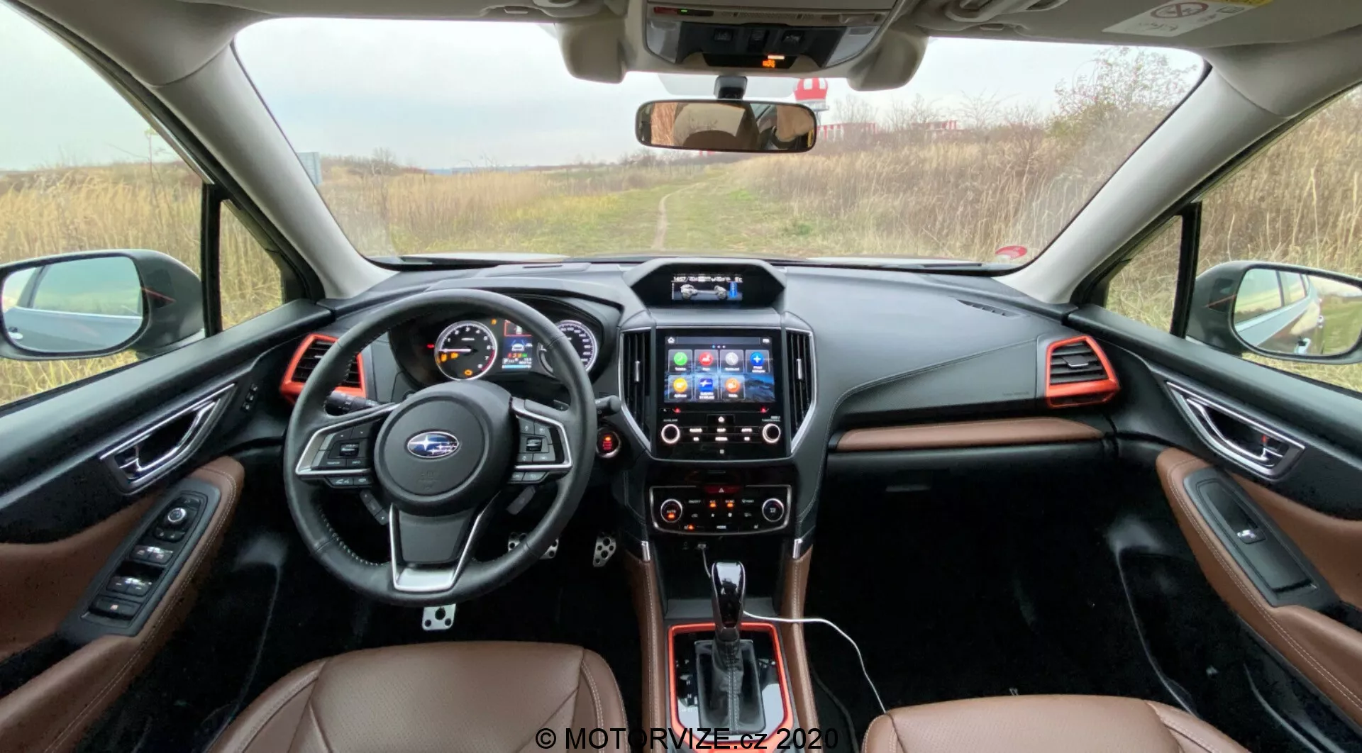 Vista interna della cabina anteriore della Subaru Forester 2020, con volante multifunzione con comandi, quadro strumenti digitale, consolle centrale con sistema di infotainment touchscreen, cambio automatico e interni bicolore con pelle marrone e rifiniture metalliche, visibile dall'area dei sedili posteriori verso il cruscotto.