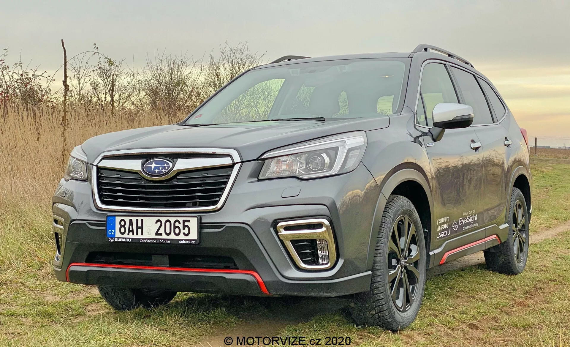 L'immagine mostra un SUV Subaru Forester 2020 di colore argento parcheggiato all'aperto durante il giorno con luce naturale. Catturata da un angolo frontale a tre quarti, la foto mostra chiaramente il lato anteriore sinistro del veicolo, inclusa la griglia esagonale con il logo del produttore, i fari LED o HID eleganti, le luci diurne a forma di C intorno ai fendinebbia e il paraurti anteriore atletico con elementi di protezione argento. Il cofano scolpito presenta creste rialzate e i passaruota sporgenti conferiscono un aspetto muscoloso. Il profilo laterale presenta una linea di carattere prominente, rivestimenti in plastica nera nella parte inferiore delle porte, cerchi in lega ricoperti di terra, portapacchi e uno specchietto laterale parzialmente visibile con indicatori di direzione integrati. Un distintivo sul parafango anteriore indica un livello di allestimento speciale o il tipo di motore. L'ambiente suggerisce un uso leggero fuori strada, evidenziato dagli schizzi di fango sui lati del veicolo.