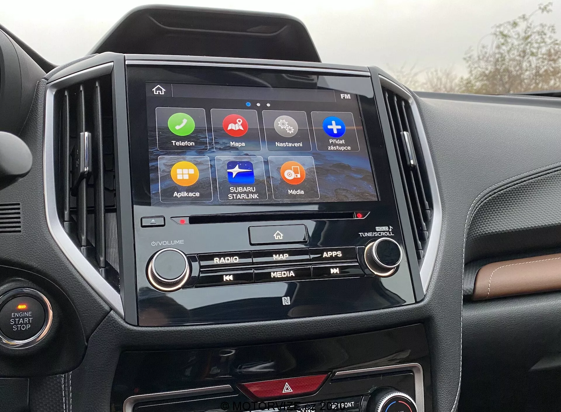 L'immagine mostra la console centrale di una Subaru Forester 2020, evidenziando il sistema di infotainment e gli interni. Al centro dell'immagine si trova lo schermo touchscreen con icone per 'Telefono', 'Mappe', 'In Riproduzione', 'Messaggi', 'Subaru Starlink', 'Media' e 'App', indicando una gamma di funzionalità come connettività, navigazione e intrattenimento. Lo schermo di infotainment è incorniciato in una cornice nera lucida che contrasta con il cruscotto bicolore. Sono visibili anche i pulsanti fisici e le manopole di controllo del clima, illustrando una combinazione di controlli digitali e manuali.