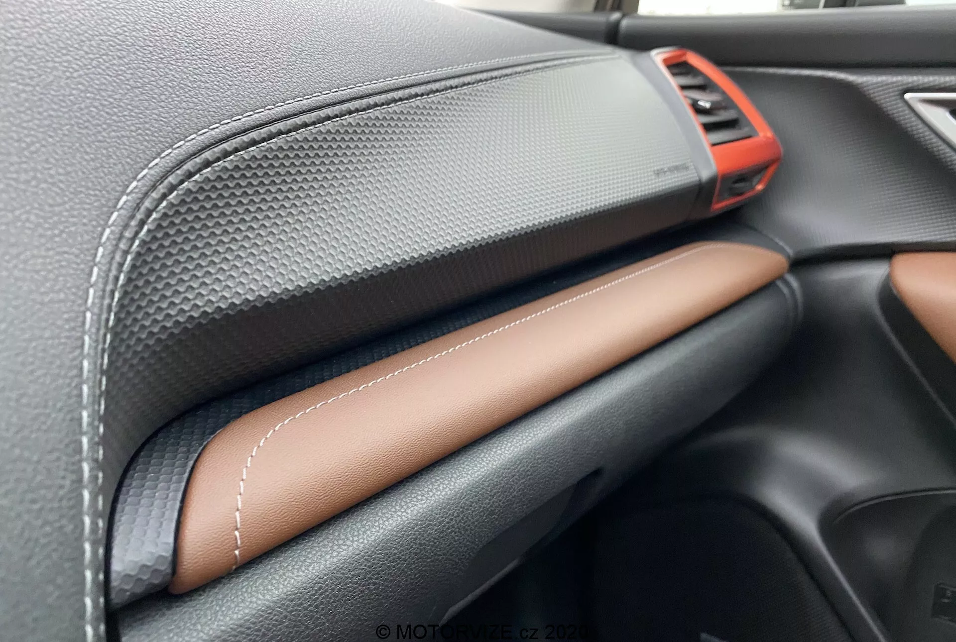 Vista ravvicinata degli interni della Subaru Forester 2020 con focus sul cruscotto. Caratteristiche includono una presa d'aria rossa metallizzata nella parte superiore destra, materiale nero con texture in fibra di carbonio sulla parte superiore del cruscotto, pelle sintetica con cuciture a contrasto in marrone chiaro o tan al centro, e un elegante vano portaoggetti nero sotto. Questo mostra l'artigianalità e la qualità dei materiali premium.