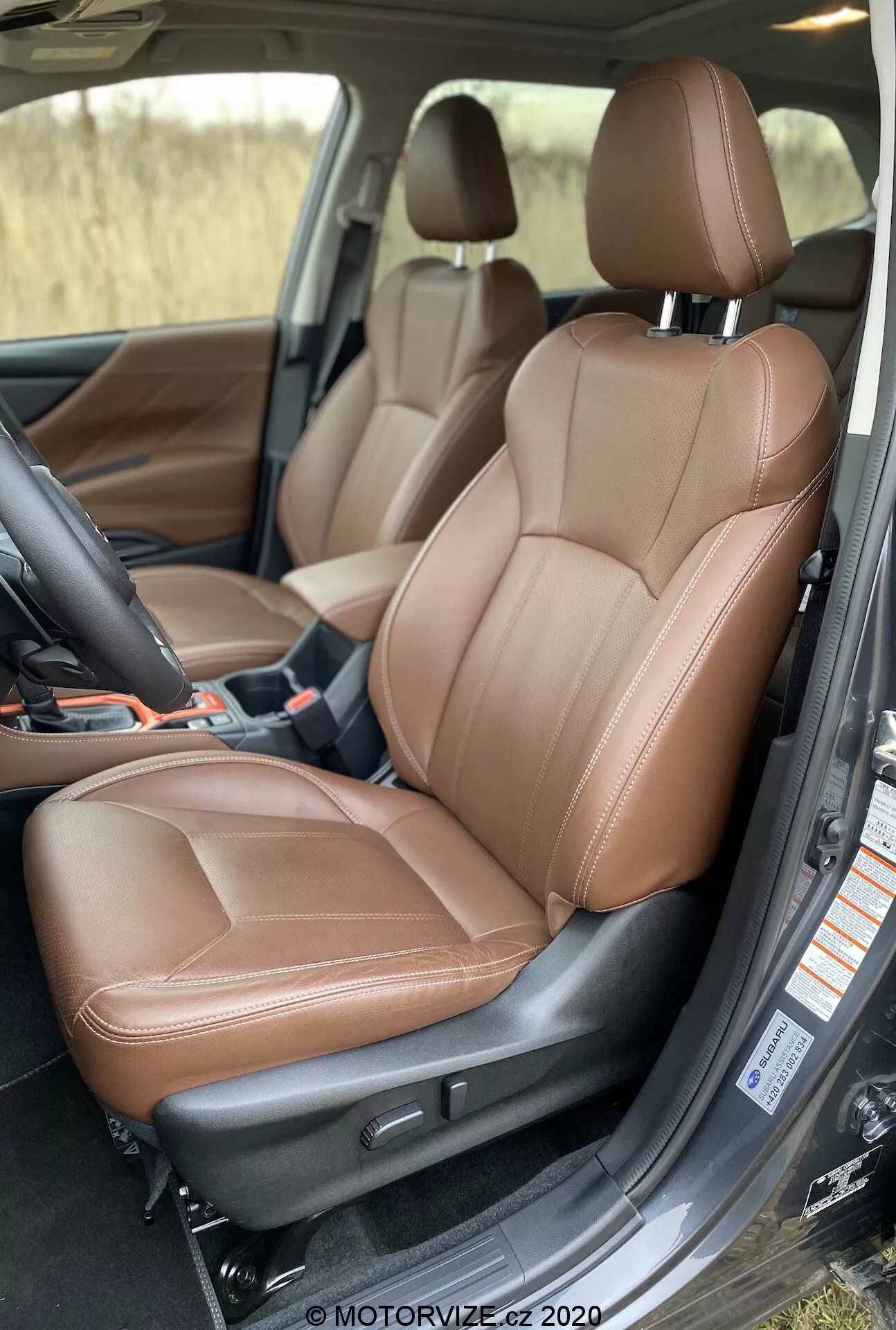 Questa immagine cattura l'interno lussuoso di una Subaru Forester 2020, focalizzandosi sul sedile anteriore del passeggero, che potrebbe anche fungere da sedile del conducente in una configurazione a guida a sinistra. Scattata da un'angolazione leggermente elevata, la foto offre una chiara visione del sedile in pelle marrone con elementi di design ergonomico, tra cui supporti laterali e supporto sagomato. Sono visibili caratteristiche come il poggiatesta regolabile, opzioni integrate potenziali di riscaldamento/raffreddamento/massaggio e funzioni regolabili elettricamente dimostrate da pulsanti e una leva di controllo simile a un joystick. La porta è aperta, rivelando parte dell'ambiente esterno, e il pannello interno della porta si allinea armoniosamente con il design del sedile. Il tappetino in moquette e la costruzione robusta suggeriscono l'alta qualità del veicolo del segmento di lusso, migliorando il comfort e la tecnologia avanzata nell'area del sedile.
