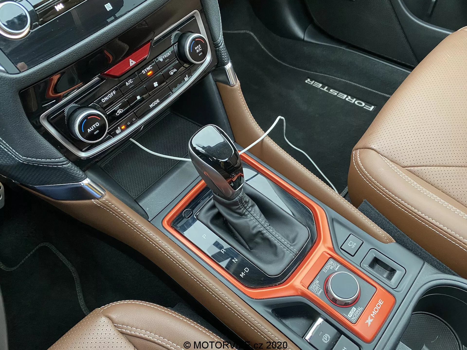 Vista interna della Subaru Forester 2020 incentrata sulla consolle centrale da una prospettiva dall'alto, con una moderna leva del cambio automatico, pannelli neri con rifiniture arancioni rossastre, interfaccia di controllo con pulsanti e manopole, pulsanti e display di controllo del clima, pulsante di avvio/arresto senza chiave, selettore di modalità di guida, interni in pelle tan con cuciture a contrasto, emblema 'BOSE' impresso sulla pelle e vista parziale del sedile passeggero e del bracciolo, evidenziando la combinazione di sportività e lusso del veicolo.