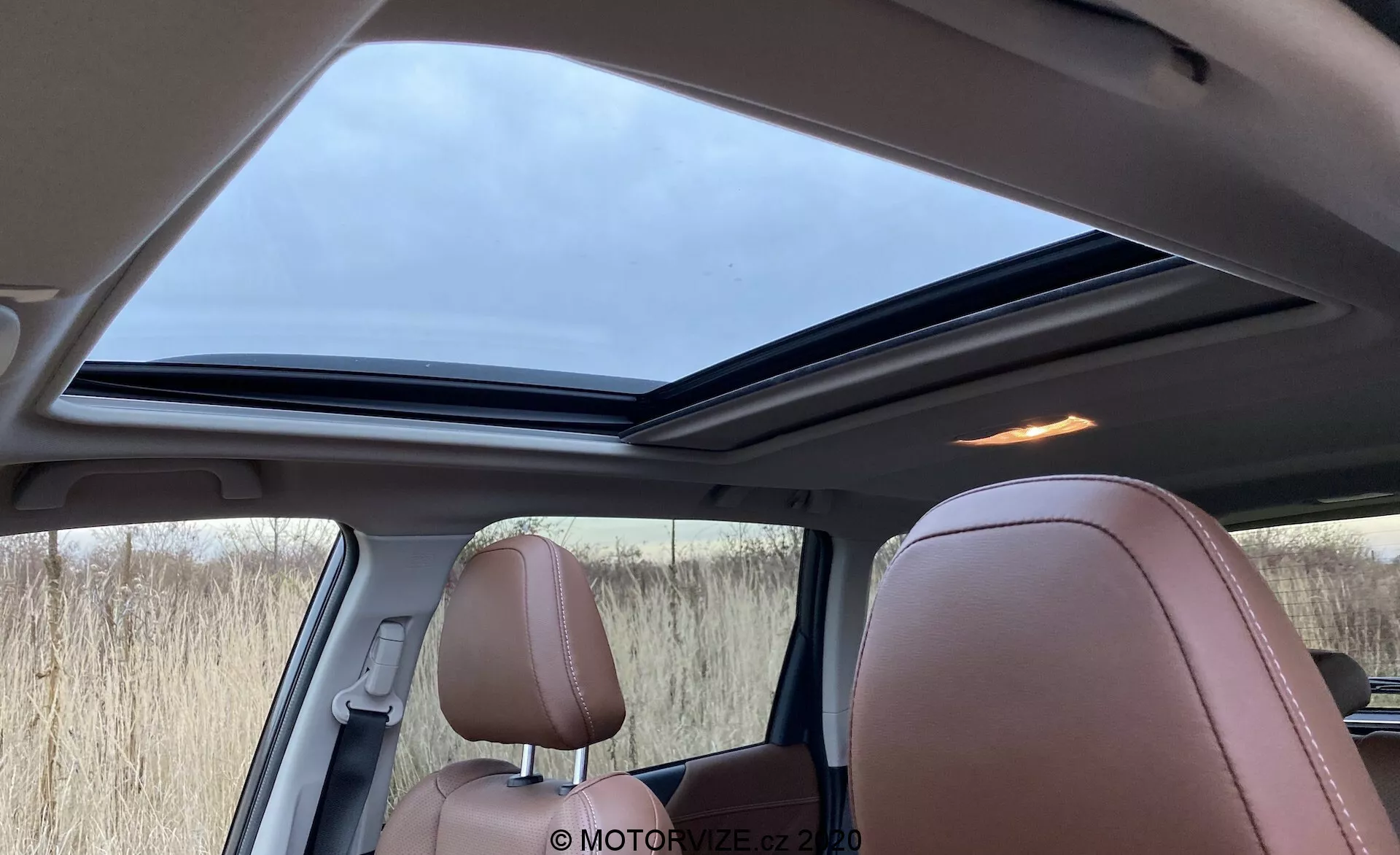 Vista interna della Subaru Forester 2020 dalla prospettiva del passeggero posteriore, con un grande tetto panoramico leggermente aperto, sedili in pelle marrone chiaro di lusso con cuciture dettagliate e una combinazione di interni marroni, neri e beige. Visibili anche una griglia per altoparlanti vicino a un pilastro e finestrini laterali oscurati, illuminati dalla luce soffusa del giorno attraverso il cielo coperto.