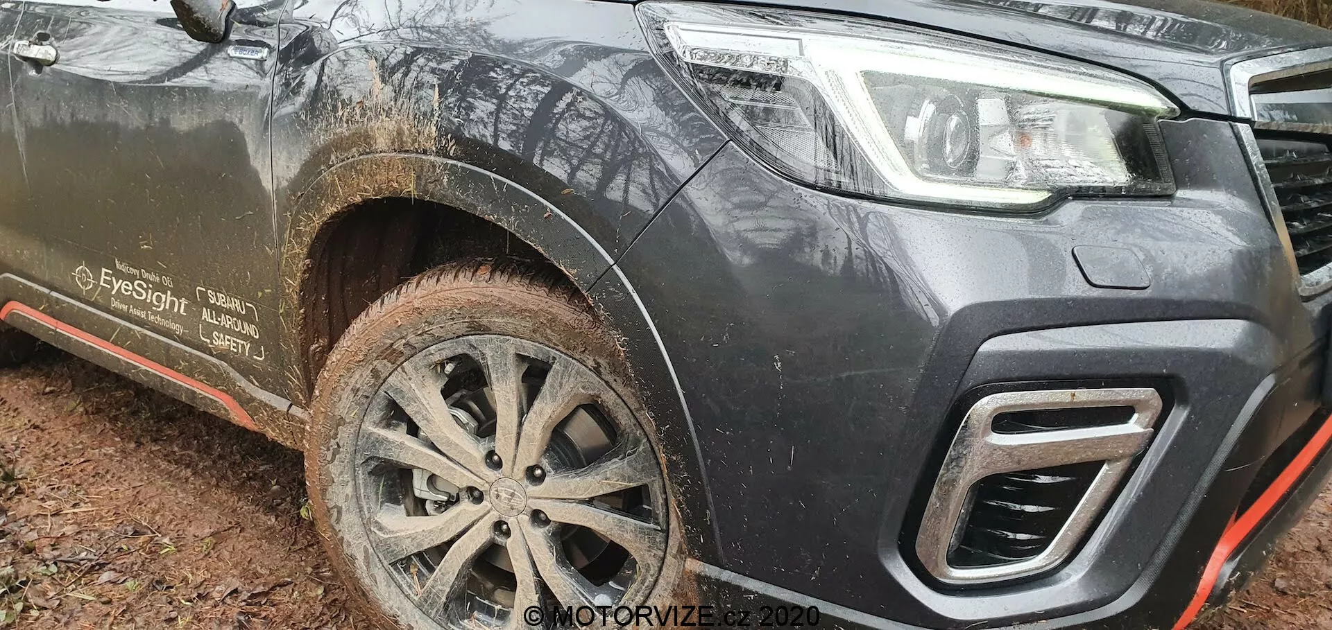 Vista del quarto anteriore sinistro di un Subaru Forester 2020 infangato, catturato da un angolo basso, che mette in risalto la sua altezza da terra, la ruota in lega multiraggio intricata, il design del paraurti inferiore e i ritagli angolari della griglia. Il veicolo di colore scuro, probabilmente nero o grigio scuro, mostra segni di utilizzo fuoristrada con schizzi di fango sulla ruota anteriore, minigonna laterale e pedana. Le caratteristiche degne di nota includono un gancio di traino integrato nel paraurti, una piastra paramotore metallica delineata in rosso per la protezione del sottoscocca e una menzione 'EyeSight', che suggerisce capacità avanzate di assistenza alla guida.