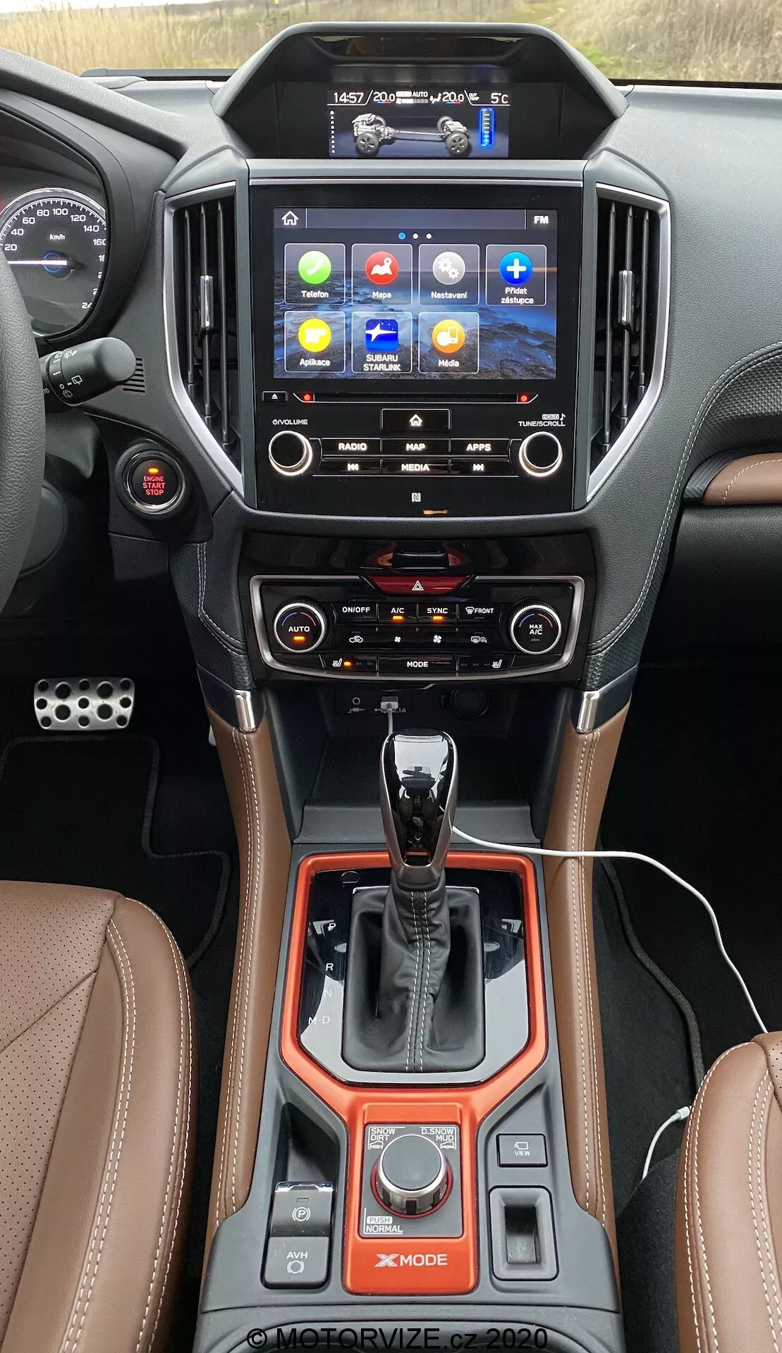 L'immagine cattura la console centrale interna di una Subaru Forester 2020 dalla prospettiva del conducente, focalizzando l'attenzione sullo stack centrale e sul tunnel. Nella parte superiore domina un touchscreen ad alta risoluzione e orientamento verticale, che mostra un colorato menu principale con icone per navigazione, telefono, media e impostazioni del veicolo. Sotto lo schermo, sono visibili due bocchette dell'aria con accenti cromati e controlli tattili per un sistema di climatizzazione a doppia zona con funzioni avanzate, inclusi pulsanti di sbrinamento. La parte inferiore evidenzia un pulsante di accensione/spegnimento, una leva del cambio automatico con finiture in pelle e cromo, selettori delle modalità di guida come 'X-MODE' e vari pulsanti per le impostazioni di guida. Un freno di stazionamento elettronico, funzione auto-hold e materiali lussuosi come pelle con cuciture a contrasto, metallo e finiture in plastica lucida migliorano la sensazione premium. L'angolazione sottolinea la tecnologia avanzata del veicolo, il comfort, la connettività e l'adattabilità.