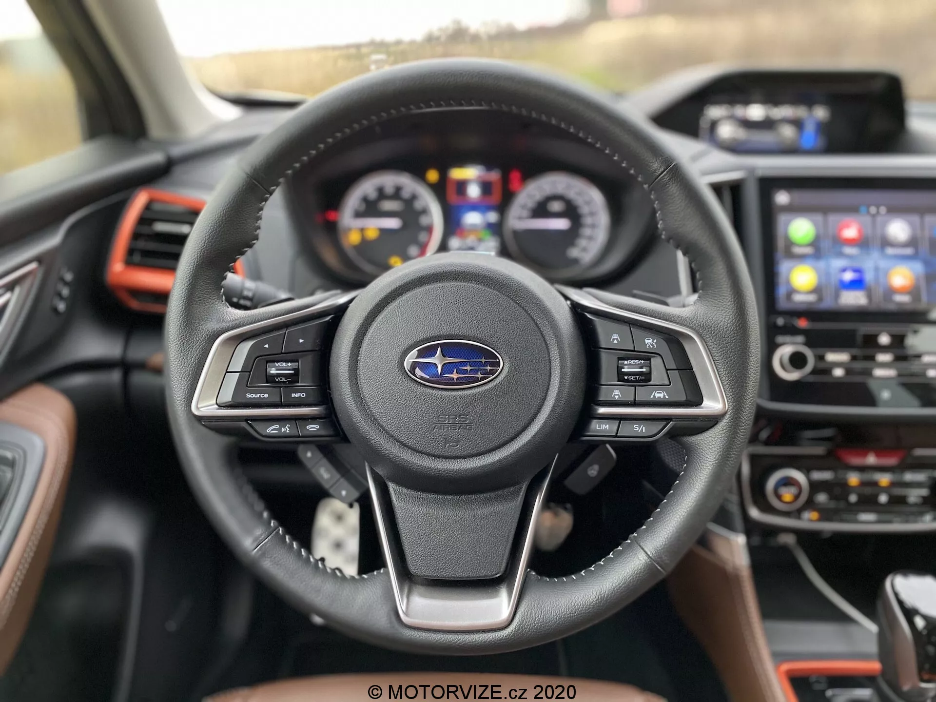 Vista ravvicinata dell'interno della Subaru Forester 2020 dal sedile del conducente, con particolare attenzione al volante a tre razze rivestito in pelle con comandi multifunzione ed emblema Subaru. Il quadro strumenti dietro include quadranti analogici e un display digitale centrale. La console centrale presenta un sistema di infotainment con touchscreen che visualizza le icone per la navigazione e la connettività del telefono. Accenti rossi sulle bocchette e sul cruscotto aggiungono un tocco sportivo all'auto moderna e ben equipaggiata.