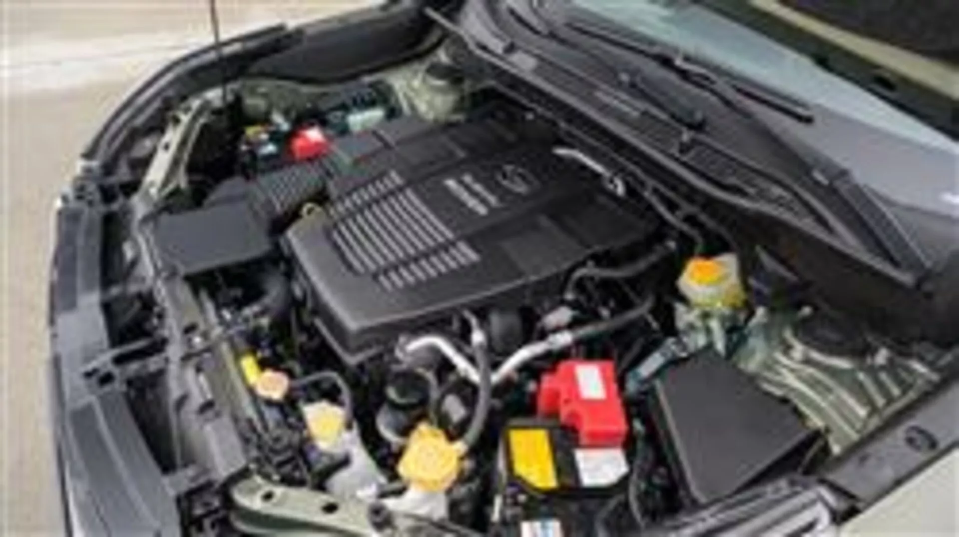 Vista ravvicinata del vano motore della Subaru Forester 2015 dal lato anteriore del guidatore mostrando la copertura del motore con il marchio, il contenitore del liquido di raffreddamento con tappo giallo e la copertura rossa del terminale positivo della batteria tra gli altri componenti meccanici ed elettrici