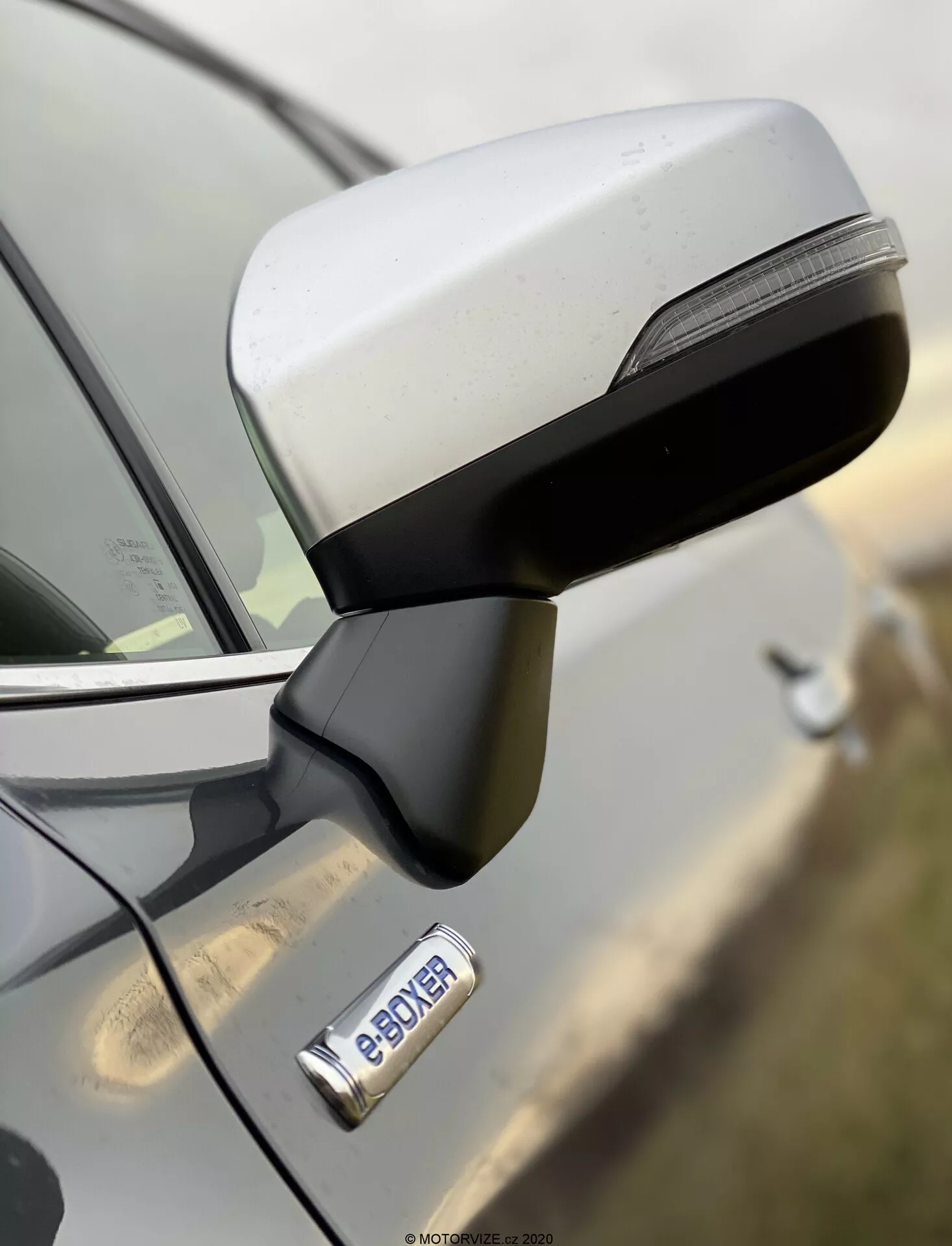 Primo piano dello specchietto laterale e della porta anteriore di una Subaru Forester 2020 da una vista laterale, evidenziando il design aerodinamico dello specchietto con indicatori di direzione a LED integrati e la maniglia della porta cromata con pulsante di accesso senza chiave. Il badge 'e-POWER' indica la motorizzazione ibrida o elettrica del veicolo. Immagine ad alta risoluzione con scarsa profondità di campo, evidenziando lo specchietto e il badge mentre lo sfondo è sfocato.