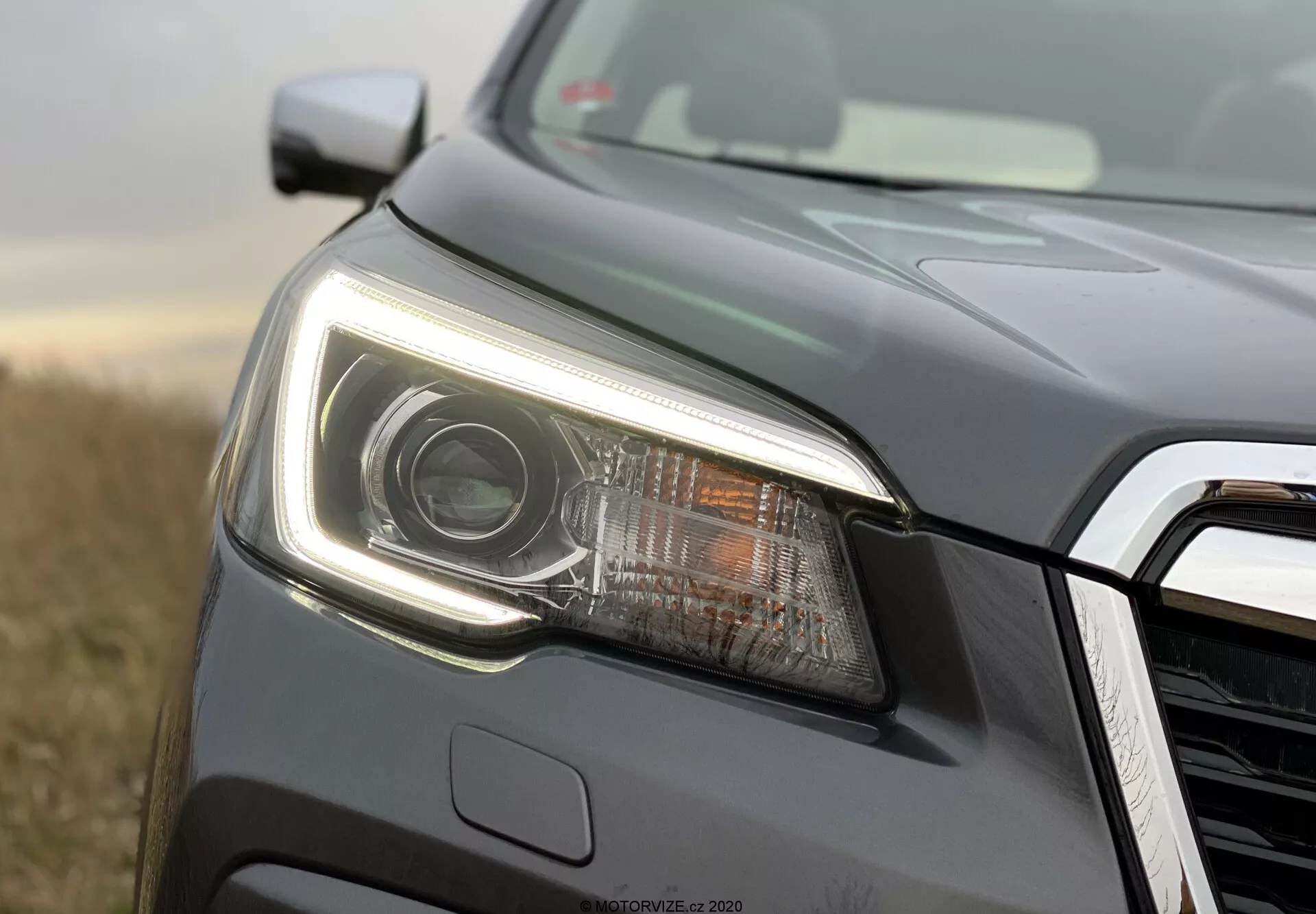 Primo piano del lato anteriore sinistro di una Subaru Forester 2020, concentrato sul gruppo ottico moderno. L'immagine, scattata da un angolo basso, presenta un faro prominente con lente a proiettore e illuminazione a LED, incluse le luci diurne affilate. La scocca del faro, elegante e con linee angolari e forma aerodinamica. Una parte della griglia del frontale con una finitura cromata o metallica è visibile. Il veicolo, verniciato in grigio scuro metallizzato, è all'aperto con luce naturale e un fondo vegetativo sfocato, che mette in evidenza i dettagli del faro e della carrozzeria con una messa a fuoco selettiva.
