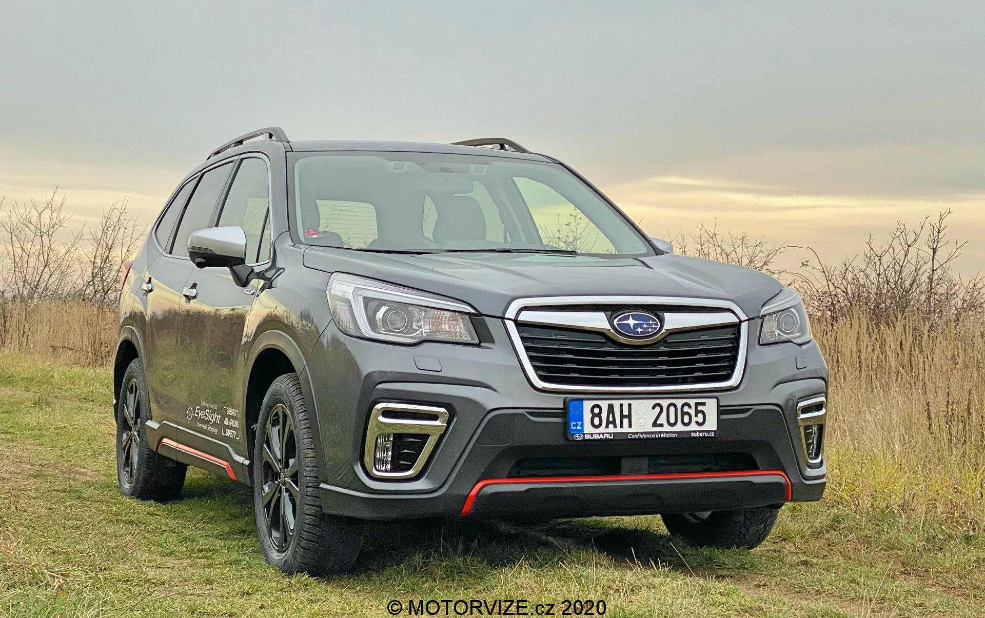 Subaru Forester 2020 nell'immagine, un SUV moderno catturato da un'angolazione frontale di tre quarti. Posizionato leggermente a destra nell'immagine su un tratto fuoristrada con erba, sotto un cielo nebbioso. L'immagine mette in risalto caratteristiche chiave come la fascia anteriore, la griglia con listello orizzontale cromato e un modulo sensore, fari LED dettagliati, linee del cofano sagomate e un profilo laterale del conducente. Cerchio in lega visibile con design multirazza, specchietti laterali con indicatori di direzione integrati, e linee della carrozzeria lungo le porte che suggeriscono dettagli di allestimento superiore. Il fango sul veicolo indica l'uso su strade sterrate e su percorsi fuoristrada. Le condizioni di luce diurna morbida e il tempo nuvoloso garantiscono un'illuminazione uniforme, mettendo in risalto il design e le capacità dell'auto.
