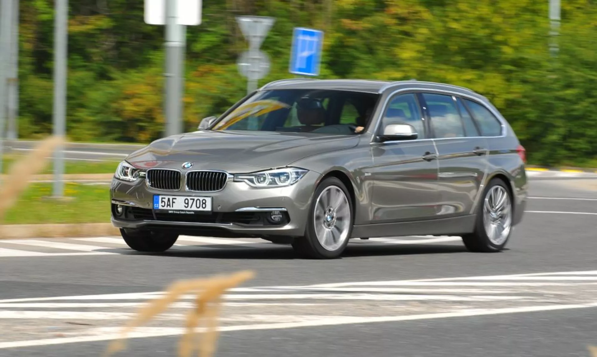 Visuale dinamica da tre quarti anteriore di una station wagon BMW argento in movimento con griglia renale distintiva e emblema, catturata da un angolo leggermente elevato che indica velocità e movimento, con focus su caratteristiche di design e tecnologia come fari angolari, ruote in lega e barre sul tetto in un contesto esterno sfocato.