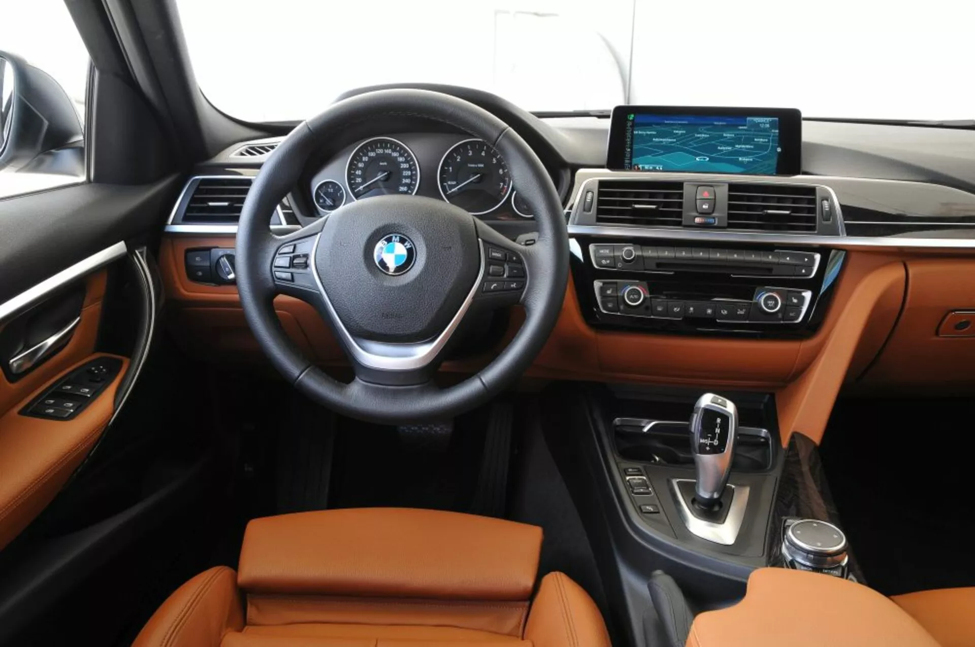Vista frontale dell'interno del BMW Outback 2003 che mostra l'area del conducente e la console centrale con comandi montati sul volante, quadranti analogici e un display digitale centrale sul cruscotto, sistema di infotainment widescreen e sedili rivestiti in pelle