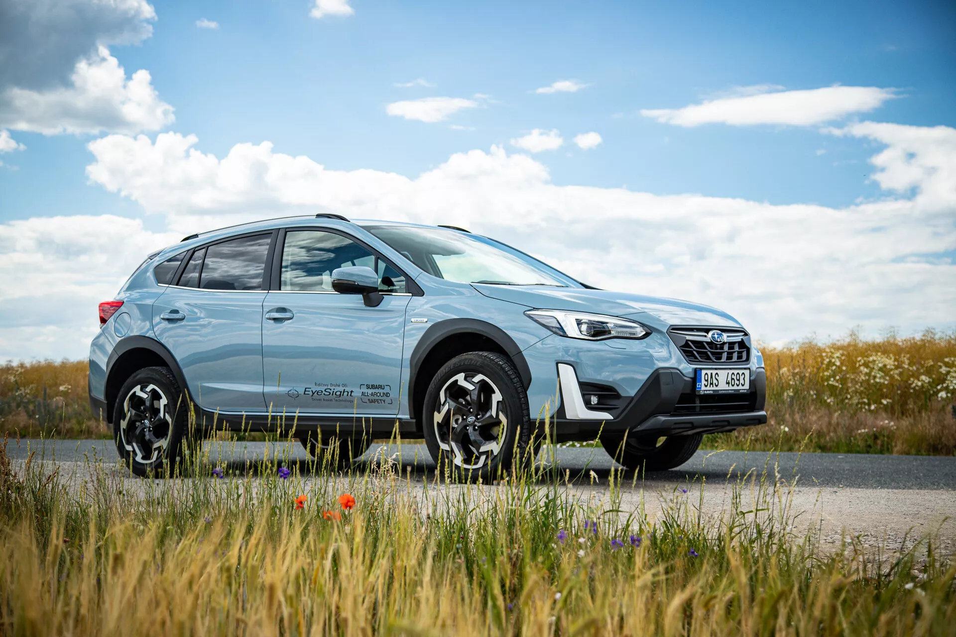 SUV crossover Subaru XV 2021 di colore azzurro chiaro parcheggiato a bordo strada con un paesaggio di campagna sullo sfondo, dotato di un design moderno con fari LED e cerchi in lega a più razze, enfatizzando le capacità sia urbane che fuoristrada.