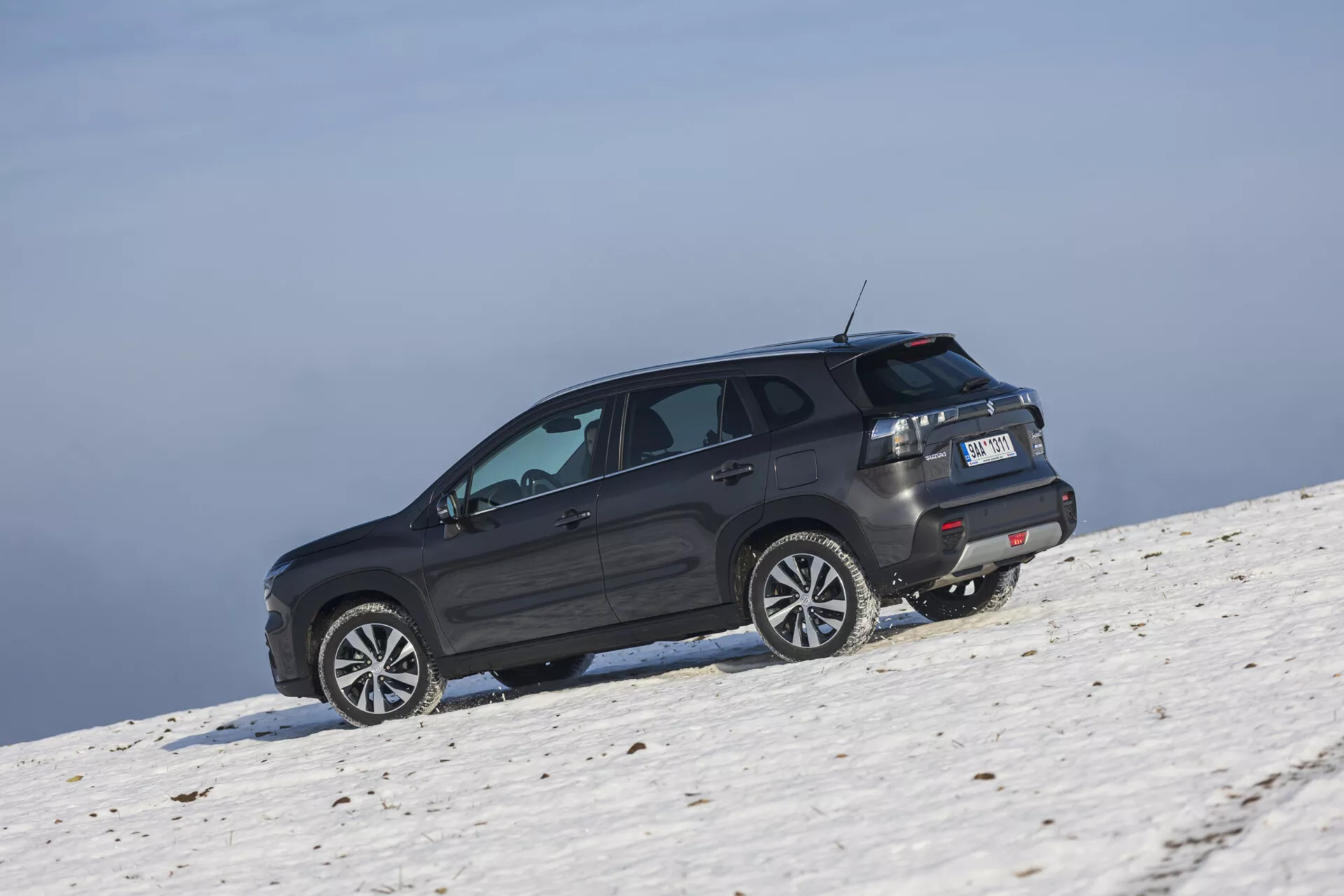 SUV Suzuki S-Cross 2022 di colore scuro posizionato su una pendenza innevata catturato da un angolo posteriore obliquo che evidenzia il suo profilo laterale e posteriore, grandi ruote in lega, finestre oscurate con sbrinatore, luci posteriori LED, spoiler sul tetto e potenziali capacità fuoristrada sotto condizioni di cielo chiaro.