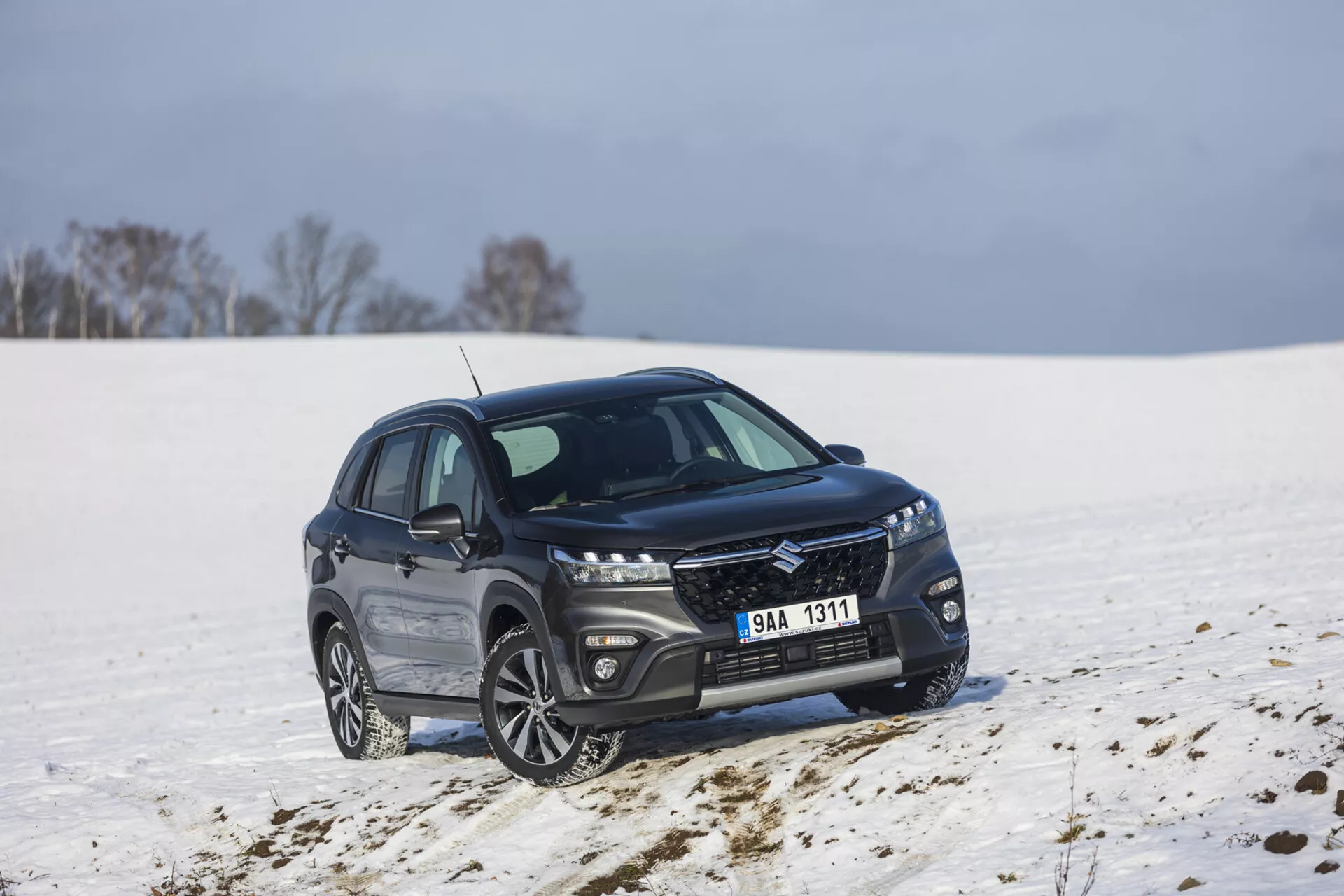 SUV compatto Suzuki S-Cross color scuro del 2022 parcheggiato in un paesaggio innevato, mostrando un profilo frontale e laterale a 45 gradi, con griglia elegante con finitura metallica, fari LED allungati, ruote in lega a più razze e profili finestre cromati lucidi che indicano un pacchetto di lusso e potenziale capacità fuoristrada