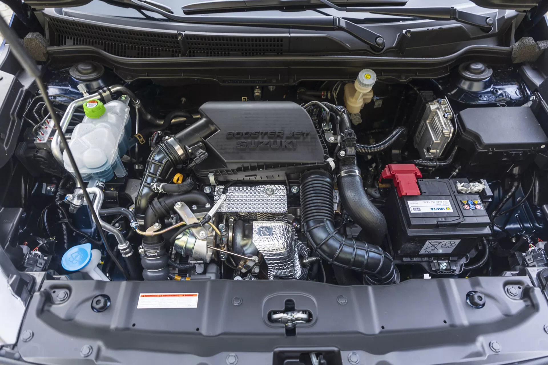 Vano motore della Suzuki S-Cross 2022 mostrato da un'angolazione frontale con componenti visibili quali il blocco motore, la testata, il collettore di aspirazione e funzionalità tecnologiche inclusi il turbocompressore e il controllo variabile delle valvole, insieme al marchio Suzuki sulla copertura del motore, in condizioni immacolate.