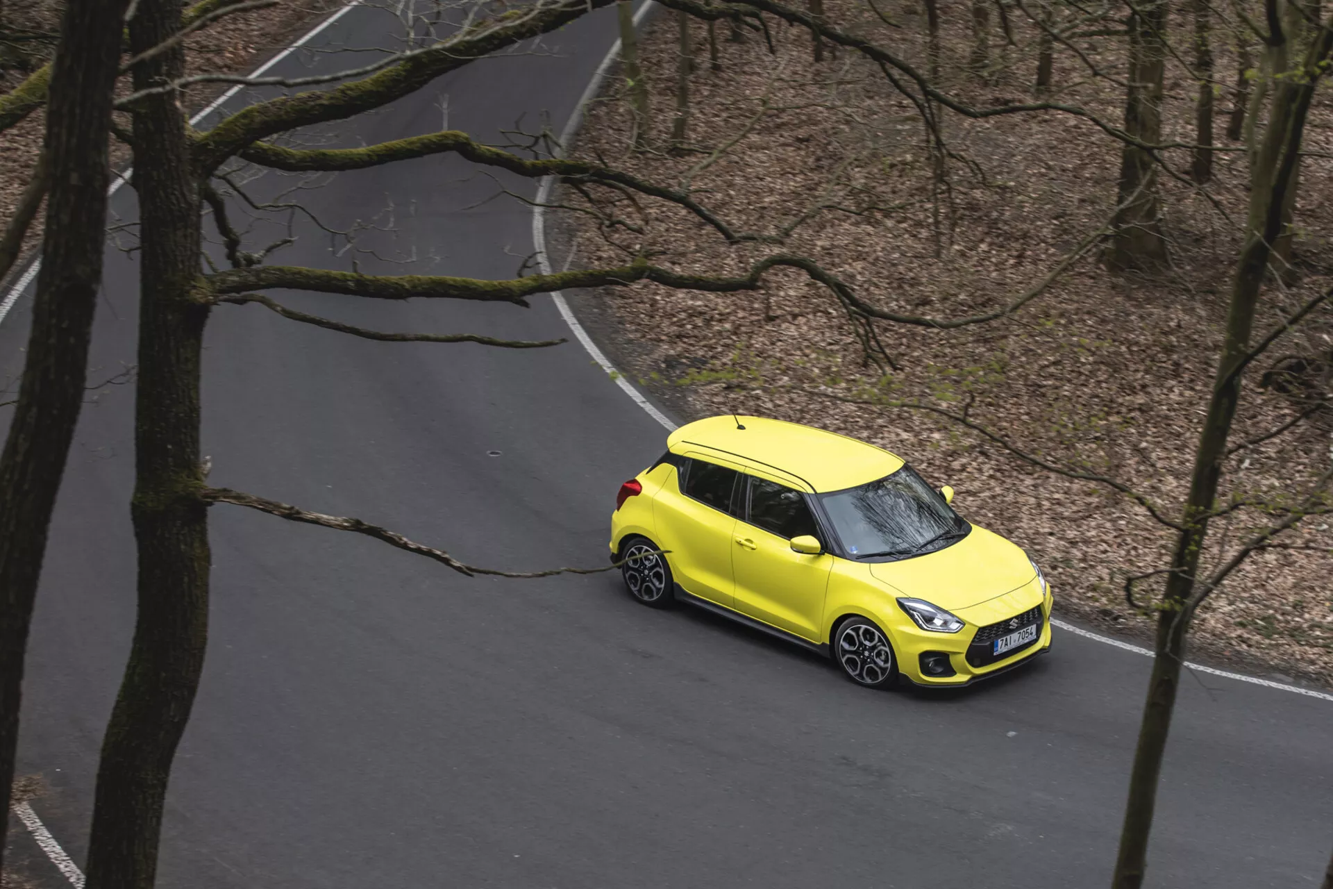 Vista aerea di una Suzuki Swift 1995 giallo brillante con una combinazione di colori a due toni che viaggia su una strada tortuosa attraverso una foresta, mostrando cerchi in lega, fari anteriori LED moderni e un design compatto a hatchback.