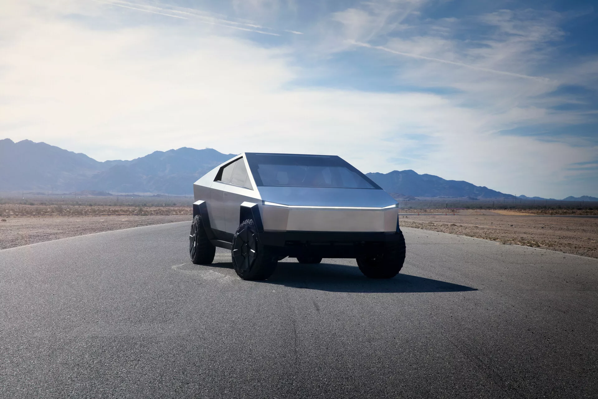 Una Tesla Cybertruck 2021 (1ª generazione) fotografata da un'angolazione frontale bassa di tre quarti, che mostra il suo design angolare e futuristico. Questa vista cattura il lato anteriore sinistro del veicolo, enfatizzando la sua forma a cuneo con linee affilate e superfici piatte. Il veicolo è dotato di grandi e robusti pneumatici adatti alla guida fuoristrada e presenta una singola striscia di luce ininterrotta lungo la larghezza del frontale, indicativa dell'illuminazione LED o laser moderna. Il parabrezza inclinato e ampio e i finestrini laterali affilati contribuiscono alla sua estetica geometrica. L'auto è realizzata in metallo spazzolato o materiale composito, suggerendo durabilità e robustezza. Lo sfondo include un cielo chiaro ed espansivo e un terreno montuoso, suggerendo un ambiente arido e desertico.