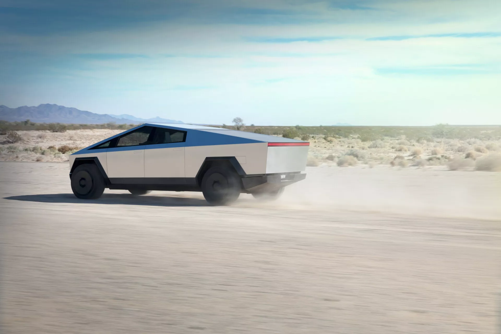 Questa immagine ritrae un Tesla Cybertruck 2021 in movimento su un terreno desertico piatto, visto da una visuale posteriore a tre quarti che mostra il lato destro e posteriore. Il camion presenta un design geometrico e angolare con un profilo a cuneo, una carrozzeria bicolore argento e grigio chiaro con una striscia a contrasto lungo la linea di cintura, e un tetto inclinato. Grandi ruote robuste con pneumatici da fuoristrada completano il design angolare. La polvere sollevata indica alta velocità. I finestrini laterali sembrano fondersi in un unico pezzo di vetro dal parabrezza al posteriore. Probabilmente un veicolo elettrico dato il design avanzato e l'assenza di tubi di scarico.
