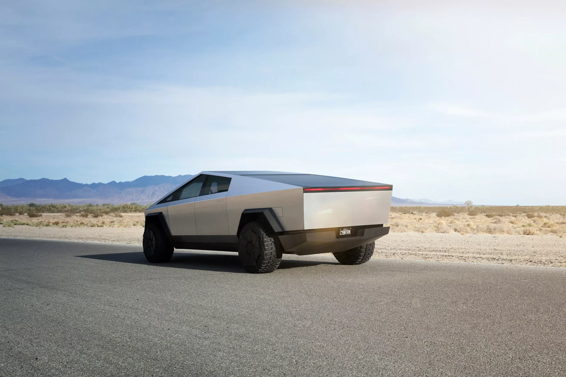 Futuristica Tesla Cybertruck 2021 su strada asfaltata piatta catturata da un angolo posteriore di tre quarti, che mette in risalto il suo caratteristico design angolare, la finitura metallica, i grandi pneumatici fuoristrada e l'assenza di specchietti e maniglie delle porte tradizionali, indicando meccanismi automatizzati avanzati.