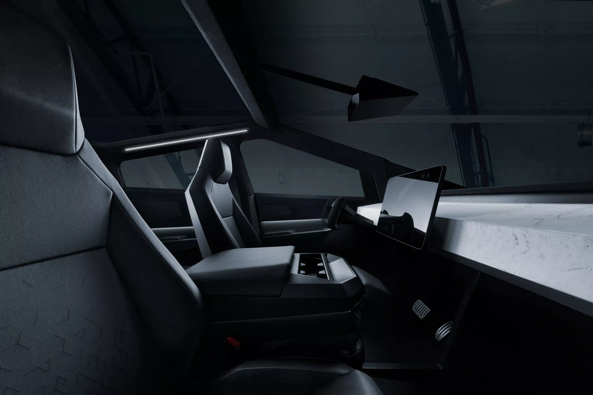 Vista interna del Tesla Cybertruck 2021 (1ª Generazione) dalla prospettiva del sedile posteriore, che mostra il lato destro della cabina con un grande display touchscreen orizzontale che domina il centro del cruscotto. Parte del volante è visibile, ma per lo più oscurata. I sedili anteriori del passeggero e del conducente presentano poggiatesta integrati e un rivestimento testurizzato, probabilmente in tessuto o pelle sintetica. Si intravede un tappetino nell'area del pavimento e non ci sono controlli manuali visibili, suggerendo un approccio minimalista e digitale. La palette di colori scuri e l'illuminazione d'accento sottile conferiscono un aspetto moderno e lussuoso.