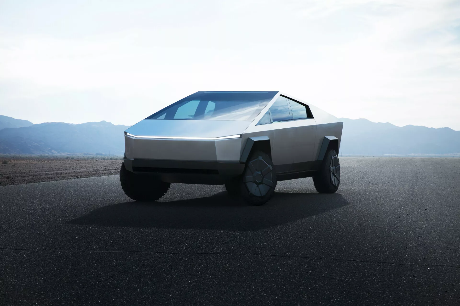 Immagine di un Tesla Cybertruck 2021 (1ª generazione) su una strada con terreni piatti o montagne desertiche sullo sfondo in pieno giorno e con cieli limpidi. La foto è scattata da un angolo basso, fornendo una vista anteriore che mette in evidenza il design angolare e futuristico del camion. Il veicolo presenta un frontale a forma di cuneo, corpo con pannelli piani, fari orizzontali sottili, parabrezza continuo inclinato, nessuno specchio laterale visibile, grandi ruote adatte per fuoristrada con cerchioni intricati e senza griglia frontale, indicando la sua propulsione elettrica.