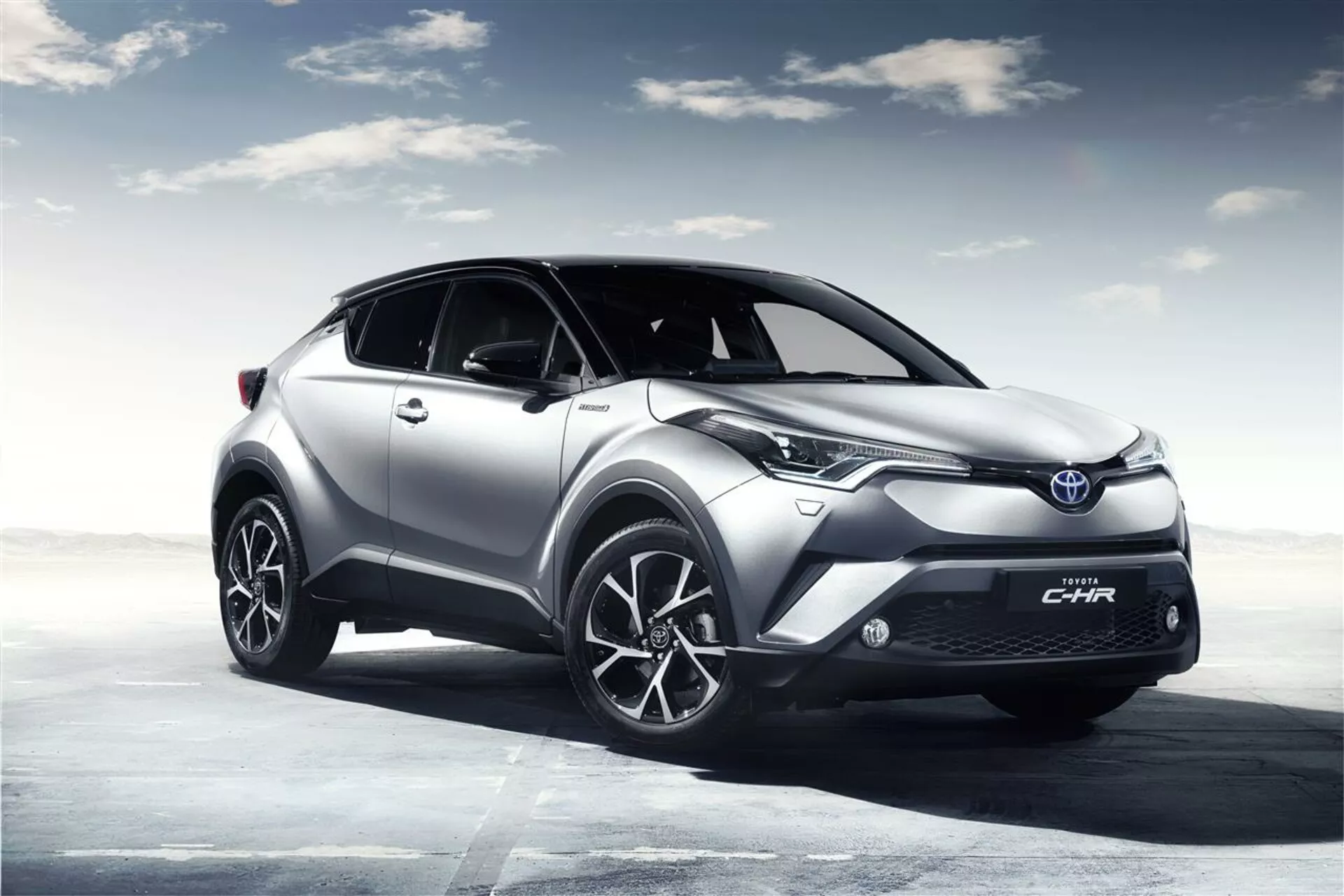 SUV crossover Toyota C-HR 2016 esposto in un paesaggio aperto renderizzato digitalmente con un cielo nuvoloso, evidenziando la sua alta distanza da terra, fari LED moderni, cerchi in lega sportivi e corpo scolpito aerodinamicamente per migliorare le prestazioni e l'efficienza.