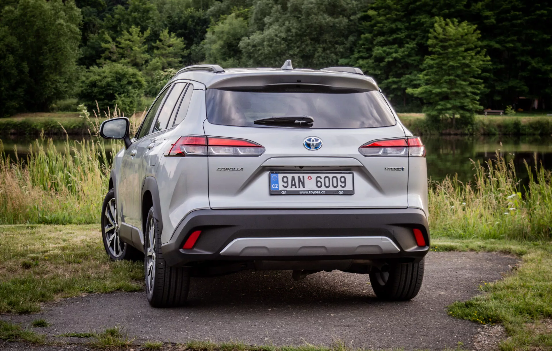 Toyota Corolla Cross argento del 2022 parcheggiata su un pavimento con verde sullo sfondo che mostra i suoi fanali posteriori LED, spoiler posteriore, ruota in lega a più razze e badge ibrido alla luce naturale del giorno