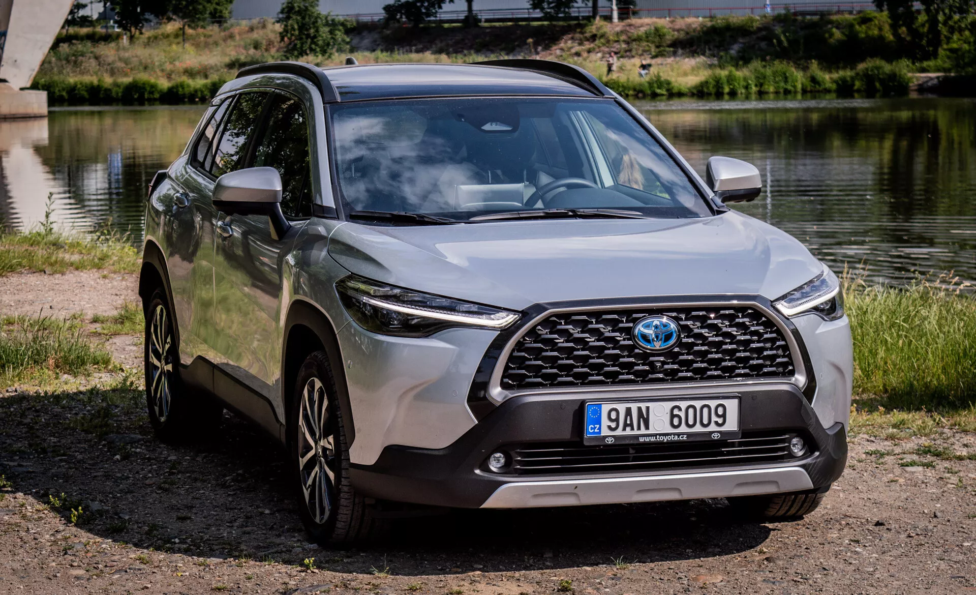 SUV crossover moderno Toyota Corolla Cross 2022 parcheggiato all'aperto vicino a un corpo d'acqua con vista chiara della sua griglia frontale, cofano, fari LED, paraurti frontale e profilo laterale compreso cerchi in lega e specchietti laterali color carrozzeria