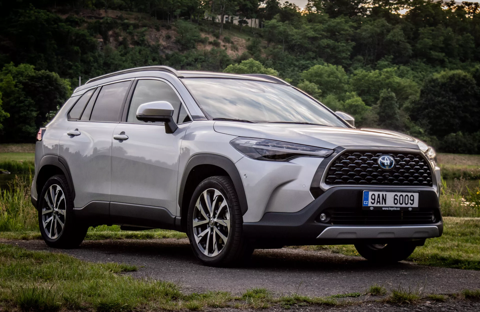 Toyota Corolla Cross 2022 in finitura metallizzata lucida che mostra il suo design moderno di SUV con griglia frontale dettagliata, emblema, fari LED angolari, paraurti anteriore aerodinamico, cofano contornato, profilo laterale con ruote in lega a più razze e barre sul tetto, scattata da un angolo basso per enfatizzare l'altezza e la postura durante il giorno in un contesto suburbano naturale.