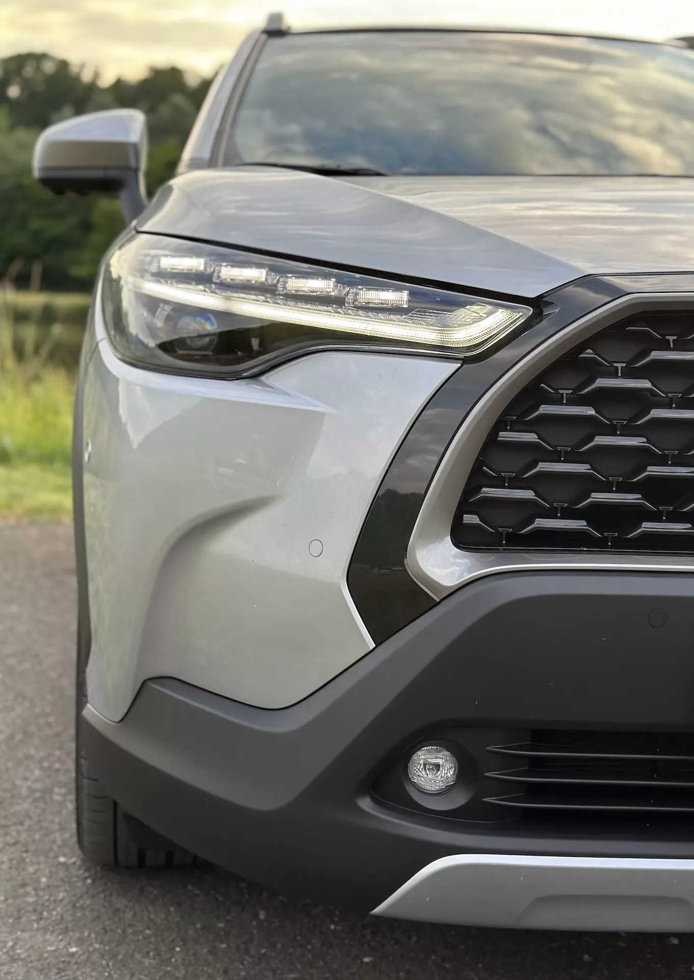 Primo piano del quarto anteriore-sinistro della Toyota Corolla Cross 2022 che evidenzia la griglia esagonale, il faro LED, la luce antinebbia, il paraurti anteriore con rivestimento protettivo, pannelli della carrozzeria eleganti, specchietto laterale con segnale di svolta integrato e finitura metallica
